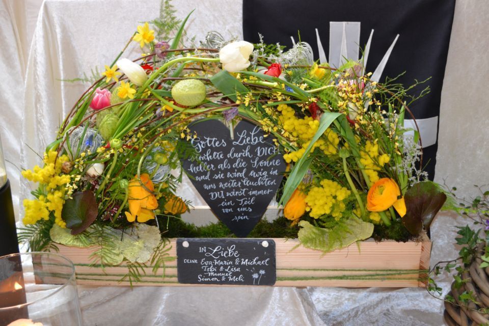 Blumengesteck in Form eines Bogens mit Gräsern, Wildblumen und Tafeln mit Beschriftungen.