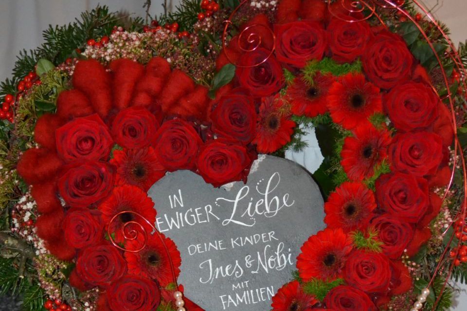 Aufgestellter Blumenkranz in Herzform mit Tannenzweigen, Schleierkraut und roten Rosen, mit einer beschrifteten Tafel in der Mitte.
