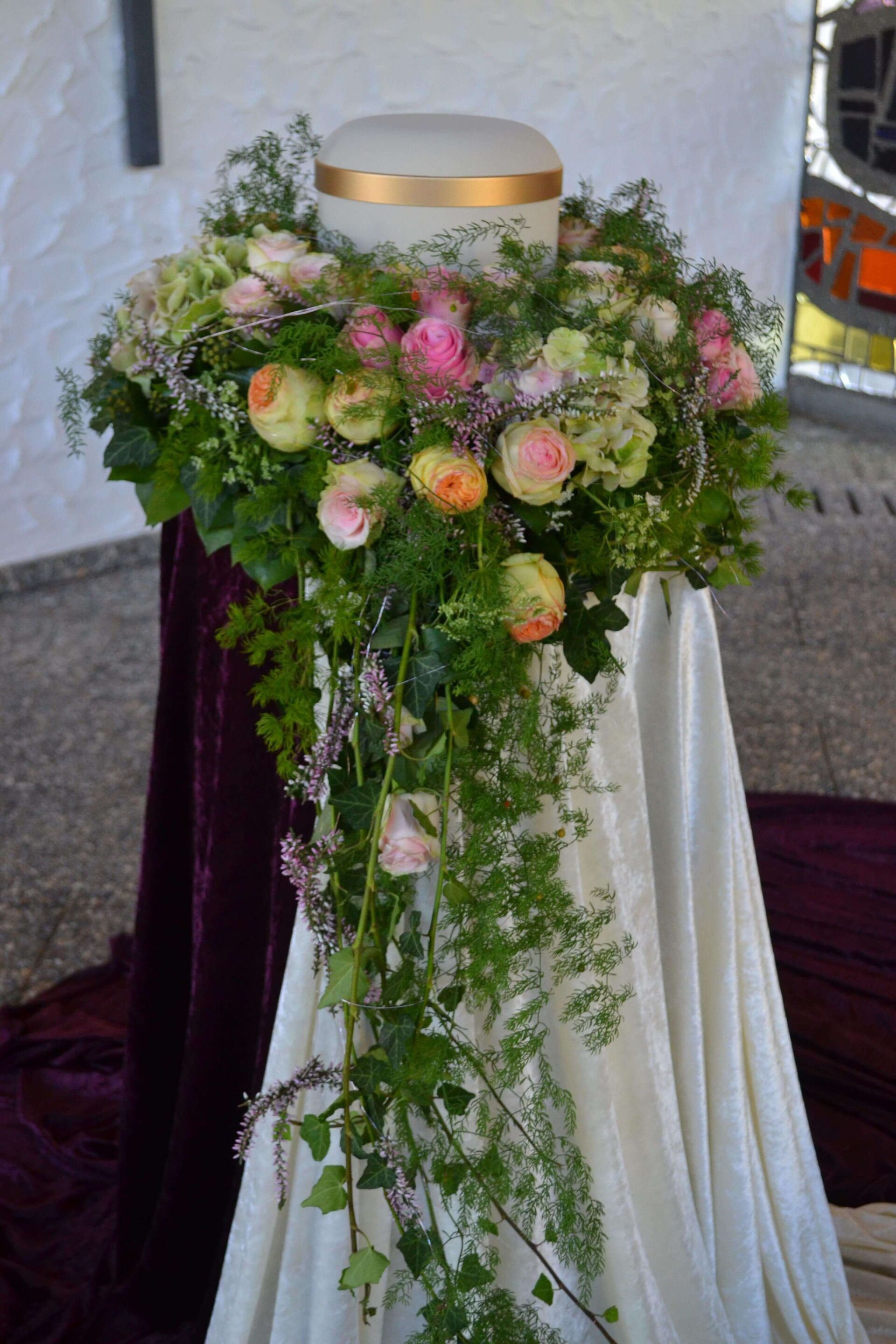 Ausgestelltes Blumengesteck um eine Urne auf einem Sockel.