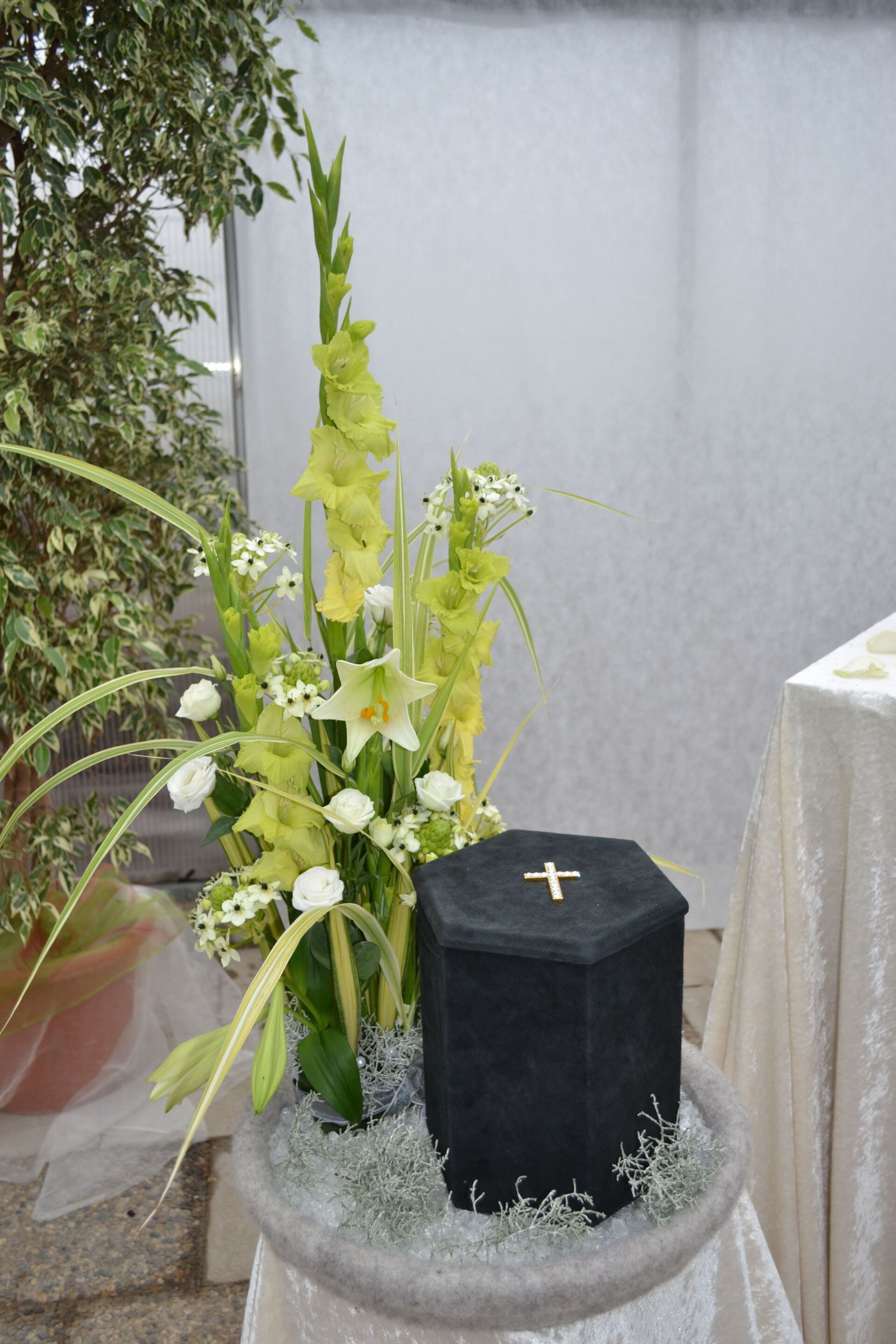 Hohe Blumengestecke um eine samtene schwarze Urne mit goldenem Kreuz