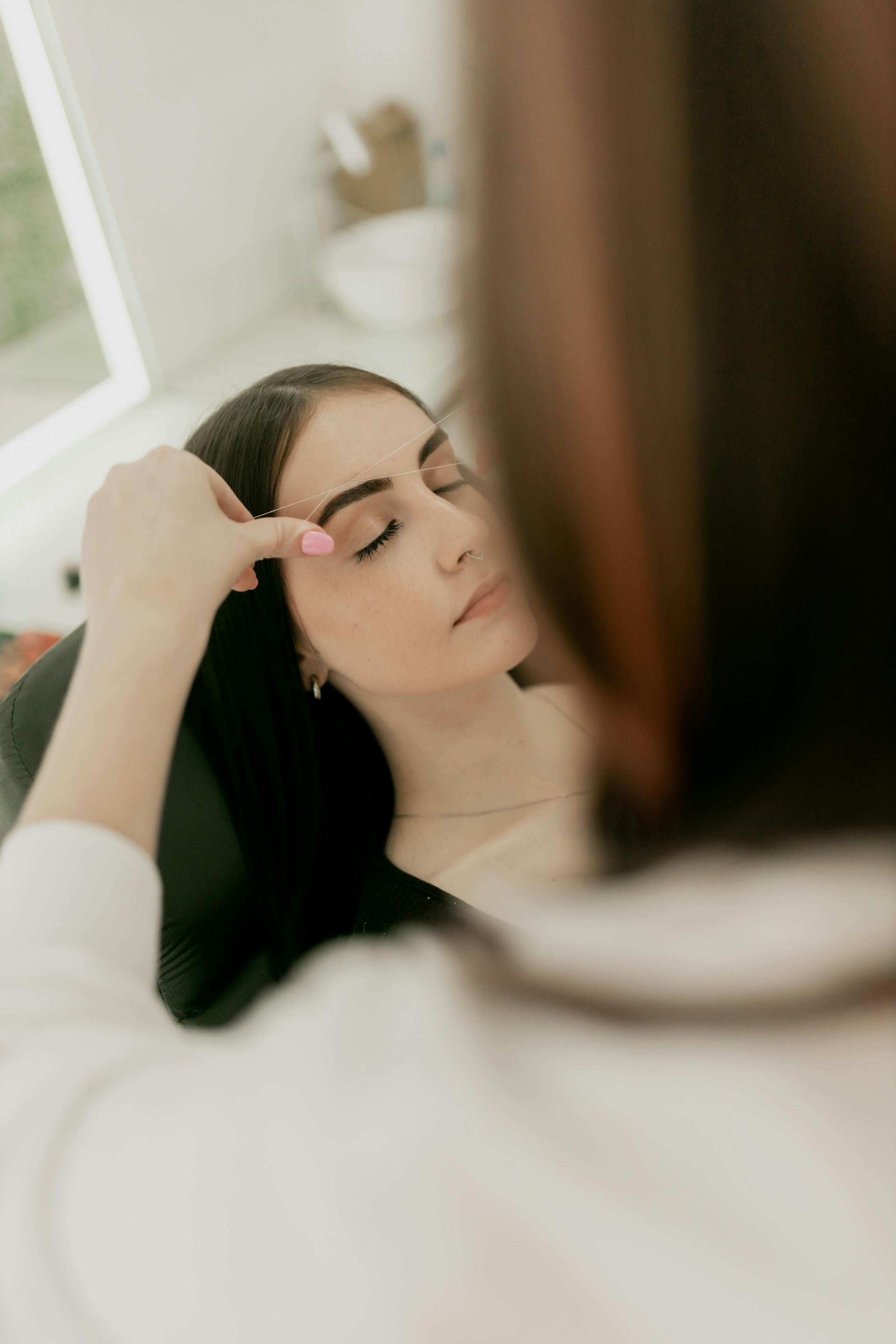 Une personne se maquille devant un miroir, utilisant un pinceau pour ses sourcils dans une pièce à la lumière tamisée.