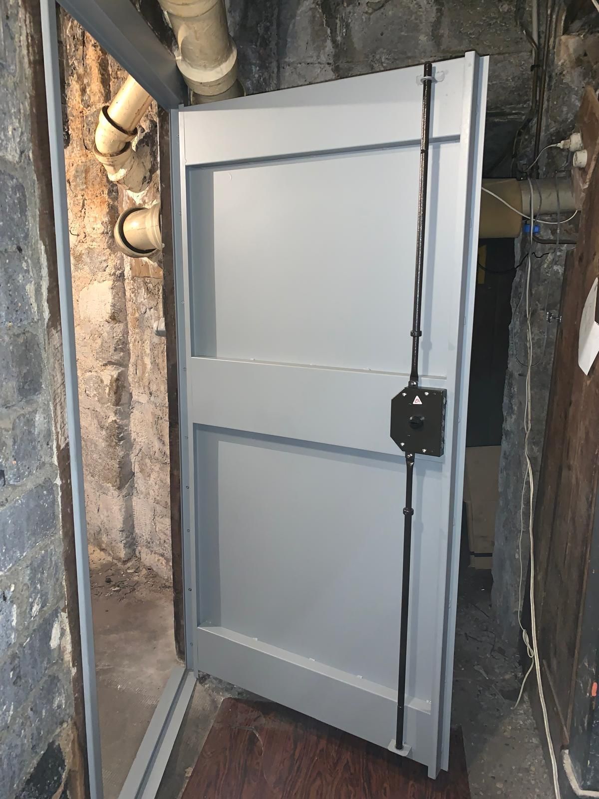 Porte utilitaire en métal gris dans un mur de sous-sol, à côté de tuyaux apparents et d'un loquet noir.