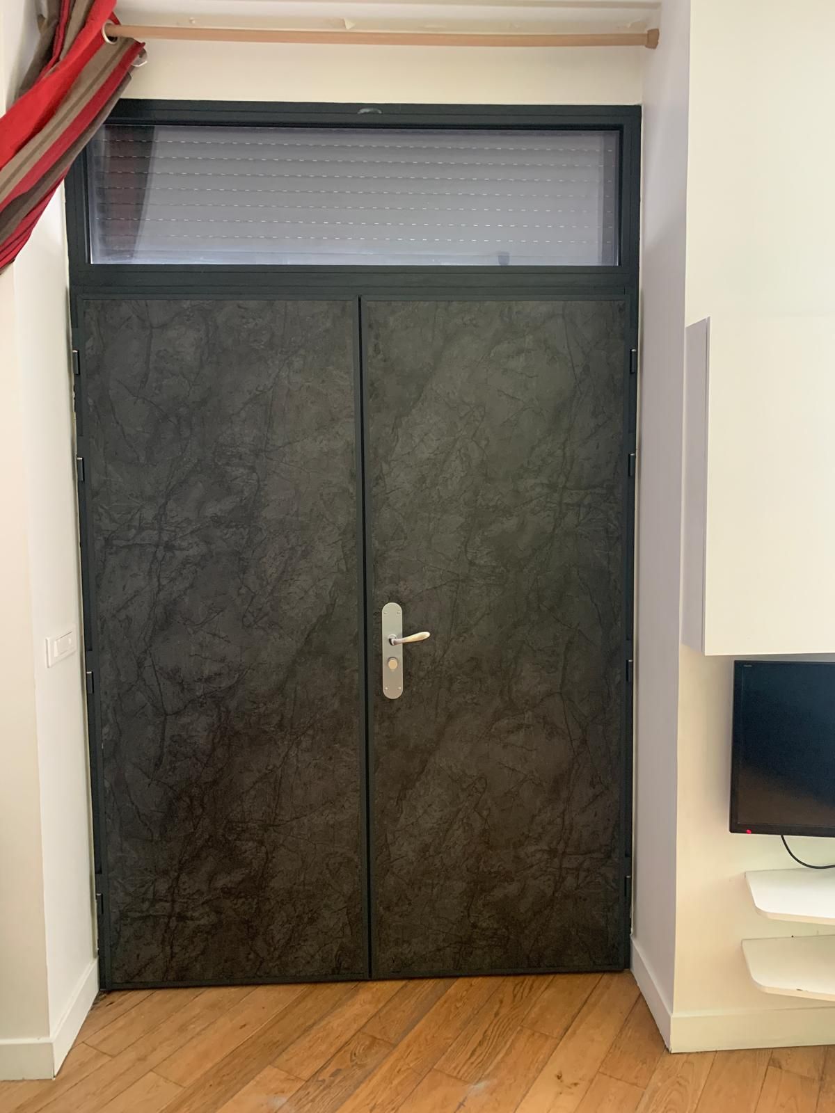 Porte de sécurité en métal gris dans un espace utilitaire étroit avec tuyauterie apparente et murs en pierre brute.