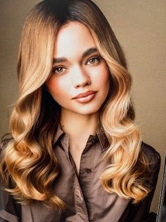 Femme aux cheveux ondulés et ombrés, portant une chemise boutonnée marron, regardant la caméra.