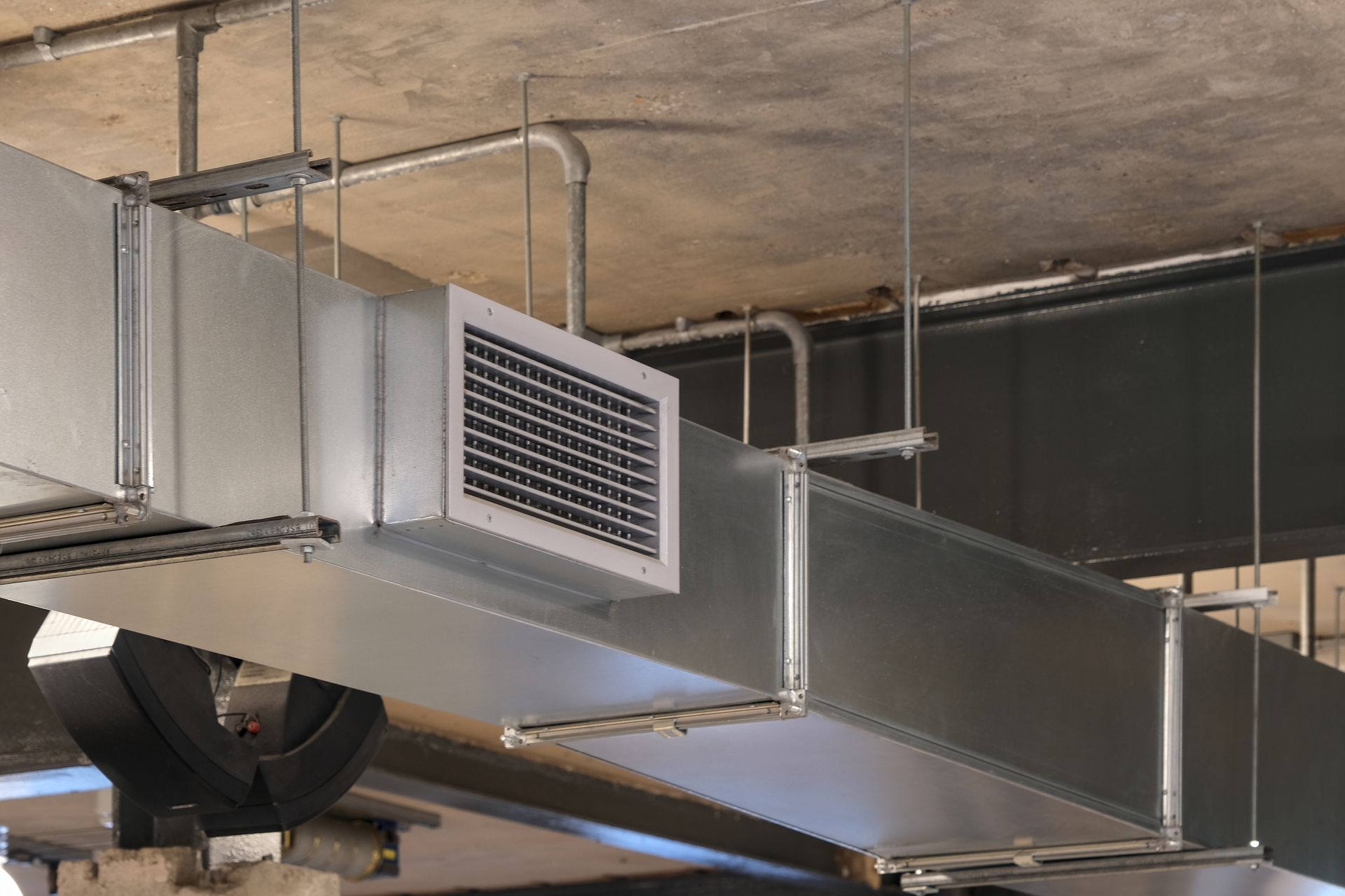 Conduits et bouches d'aération métalliques de climatisation suspendus à un plafond en béton brut à l'intérieur.