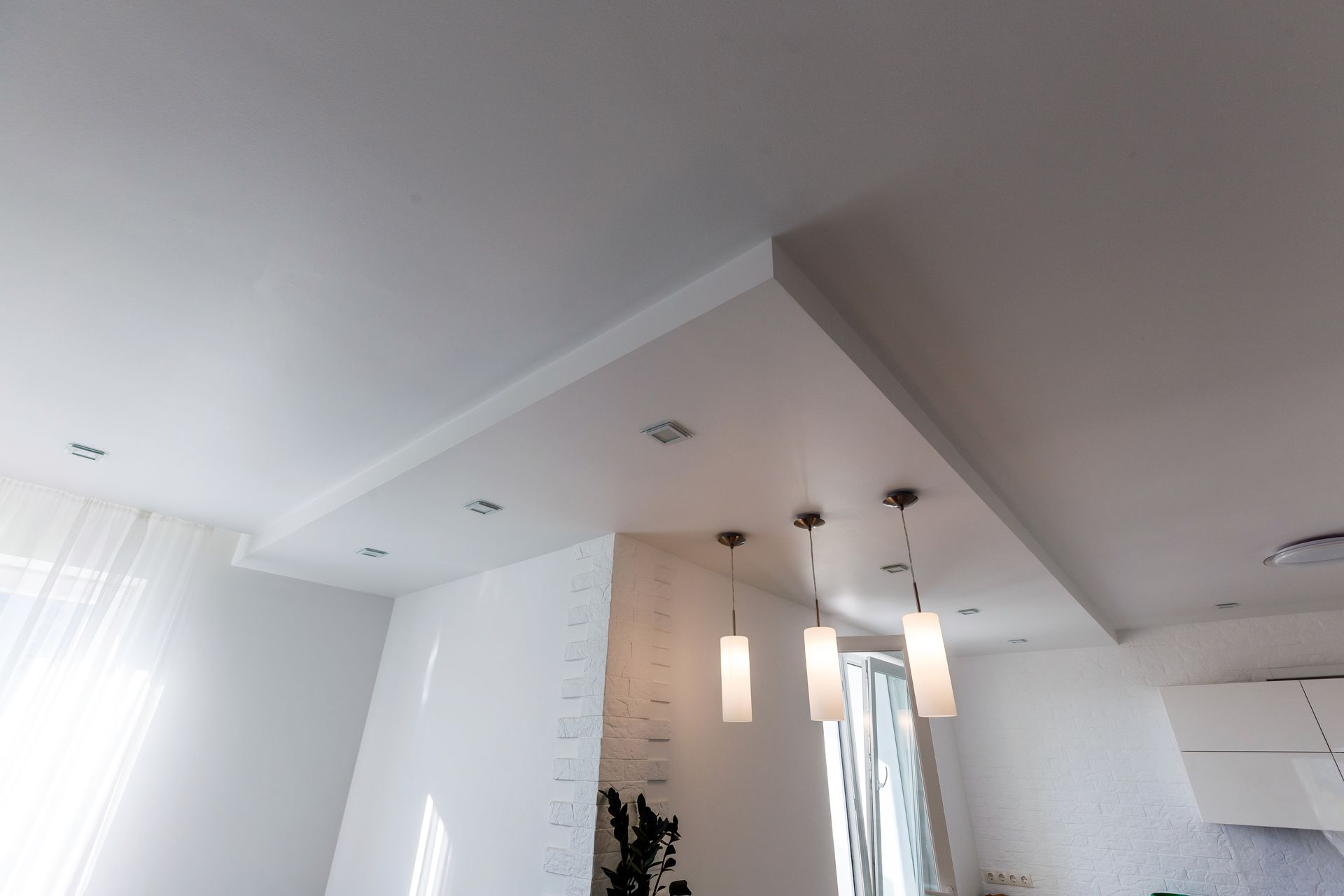 Plafond lumineux et moderne dans un salon avec spots encastrés et suspensions, murs et rideaux blancs.