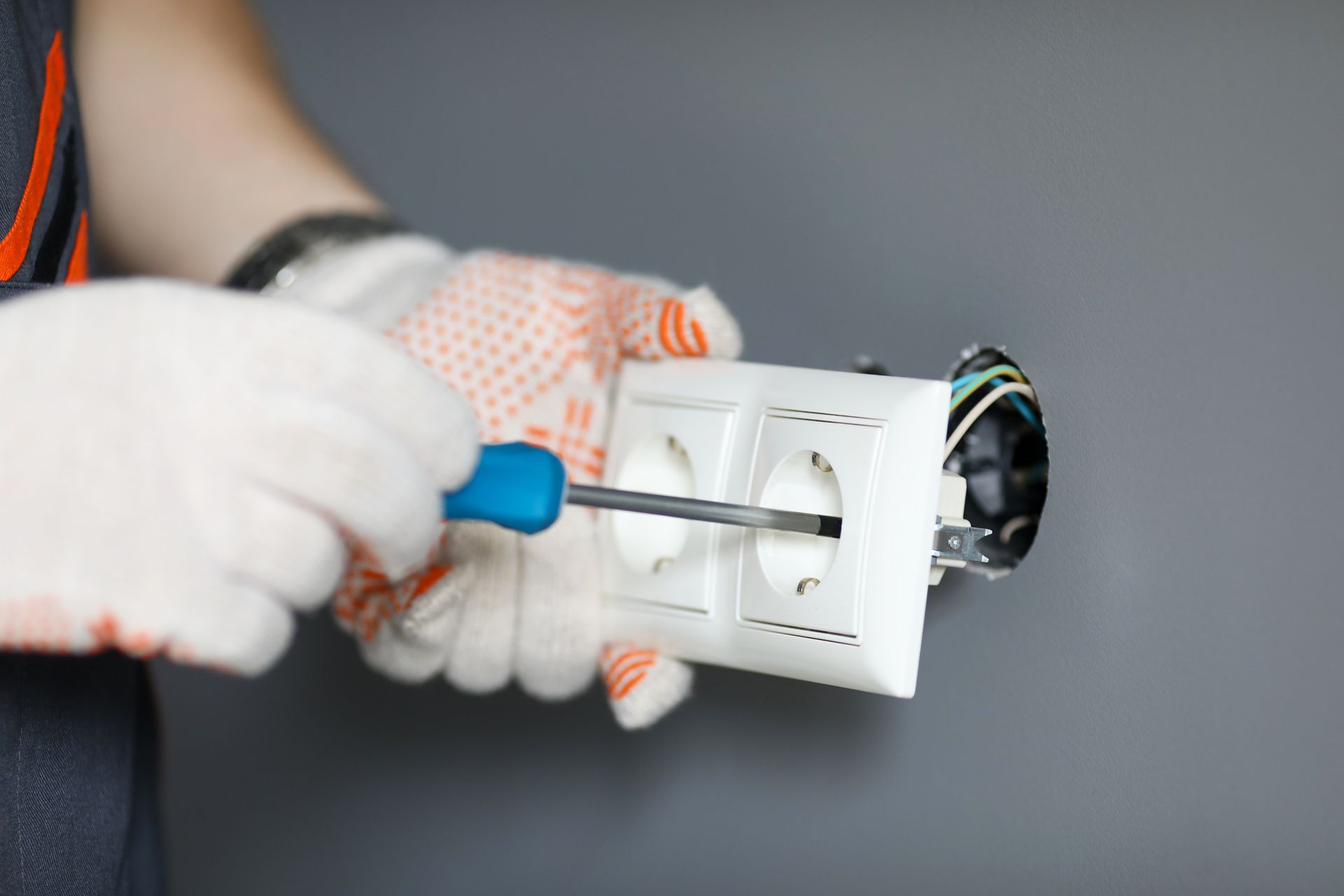 Des mains gantées utilisent un tournevis pour installer une boîte de prise électrique murale blanche.