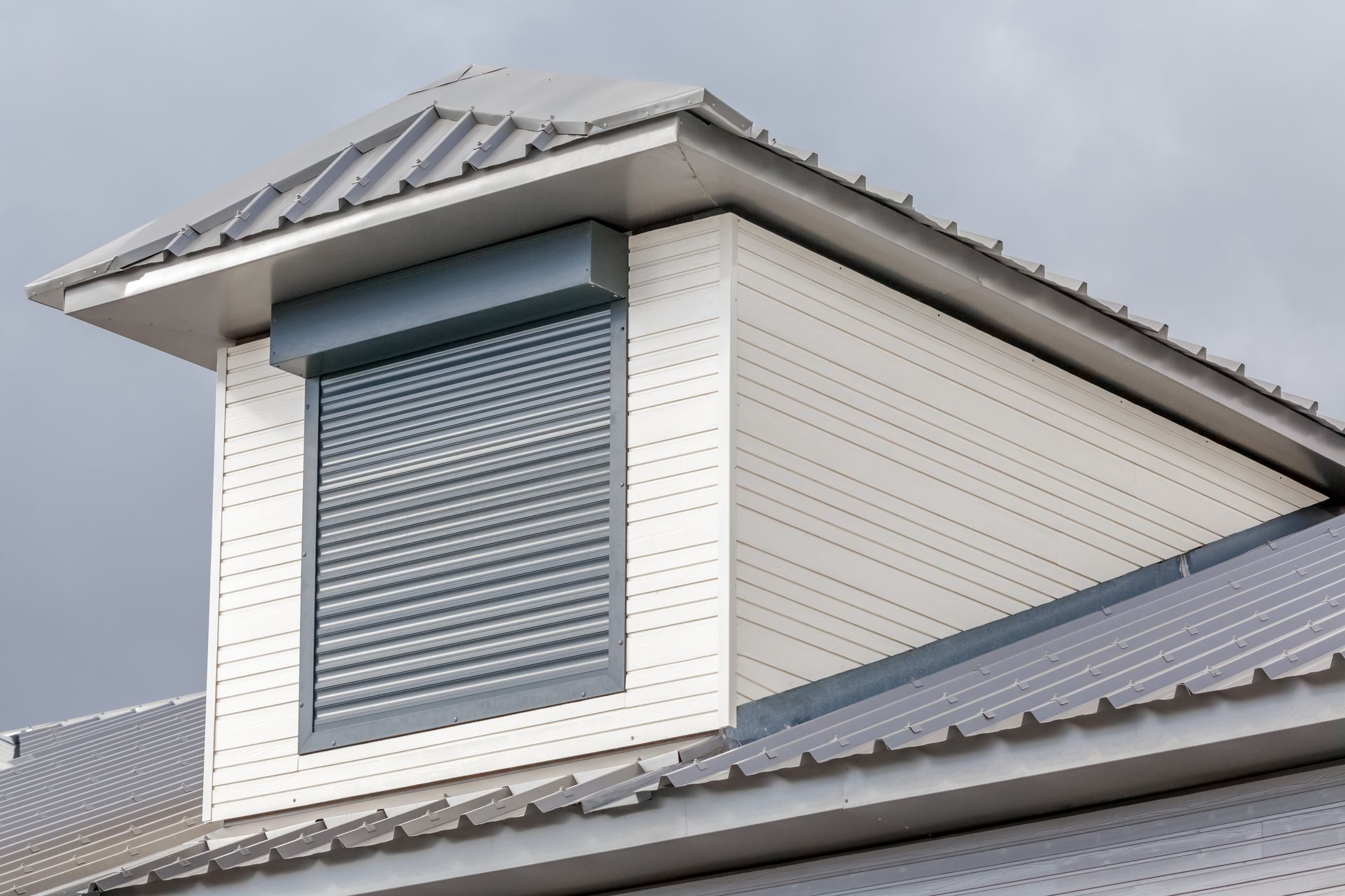 Lucarne blanche avec un volet bleu-gris fermé sur une maison à toit métallique.