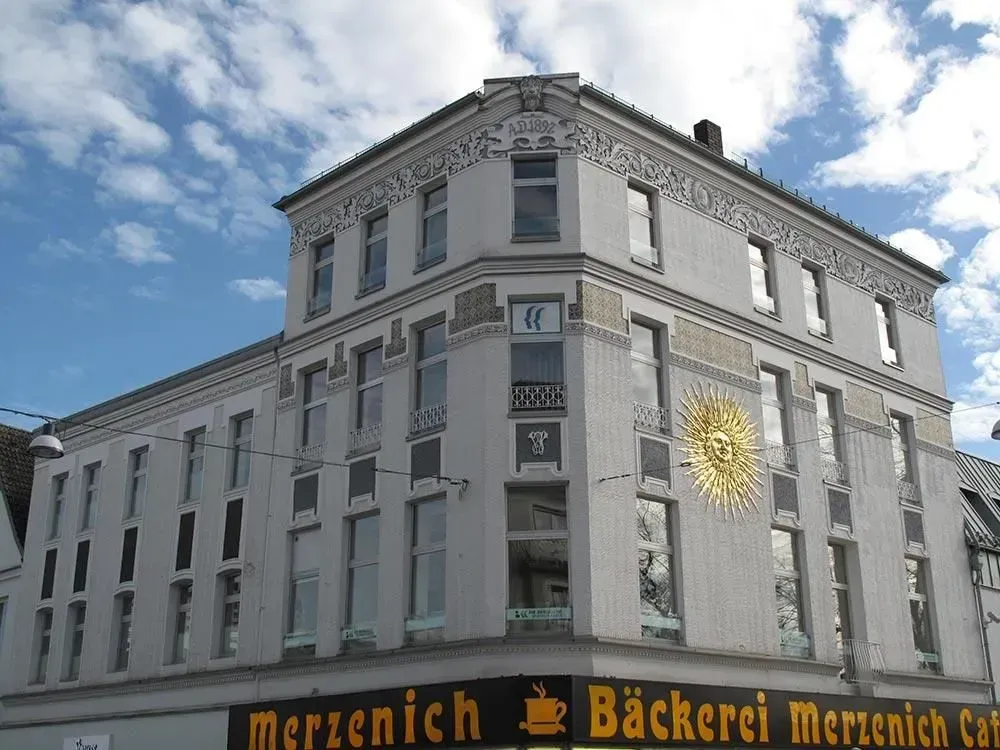 Graues Eckgebäude mit goldenem Emblem und schwarzem Ladenschild mit der Aufschrift „Menzenc“; blauer Himmel darüber.