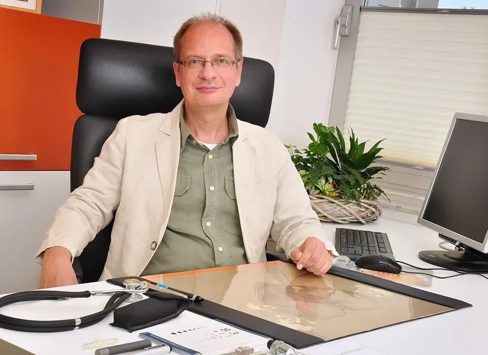 Ein Mann sitzt an einem Schreibtisch mit Papieren, einer Tastatur und einem Computermonitor neben einer Topfpflanze.