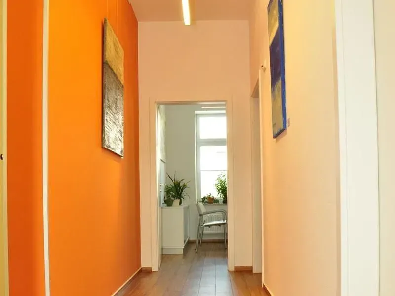 Schmaler Flur mit orangefarbenen Wänden, weißen Türen und einem lichtdurchfluteten Zimmer am Ende