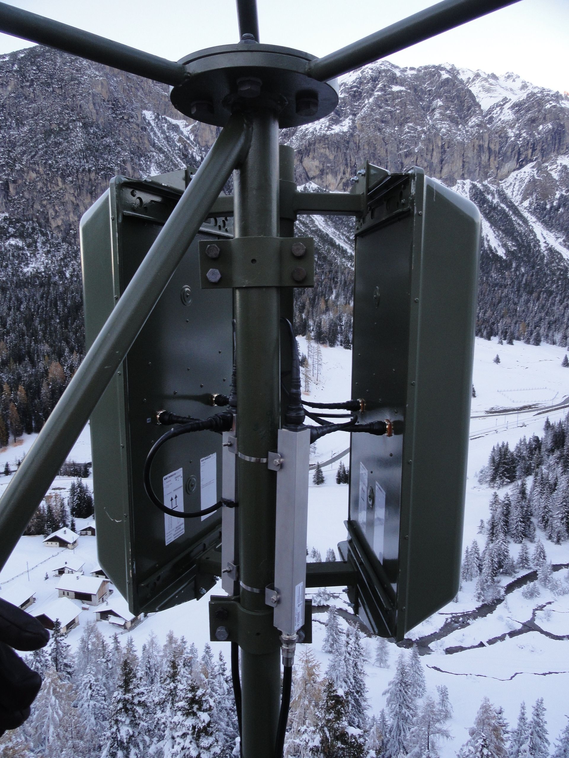 Metallener Antennenmast an der Seite eines Gebäudes an einem Berghang, umgeben von Landschaft.