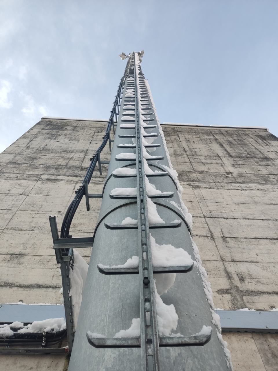 Eine an einer Betonwand befestigte, schneebedeckte Metallleiter führt hinauf in einen bewölkten Himmel.