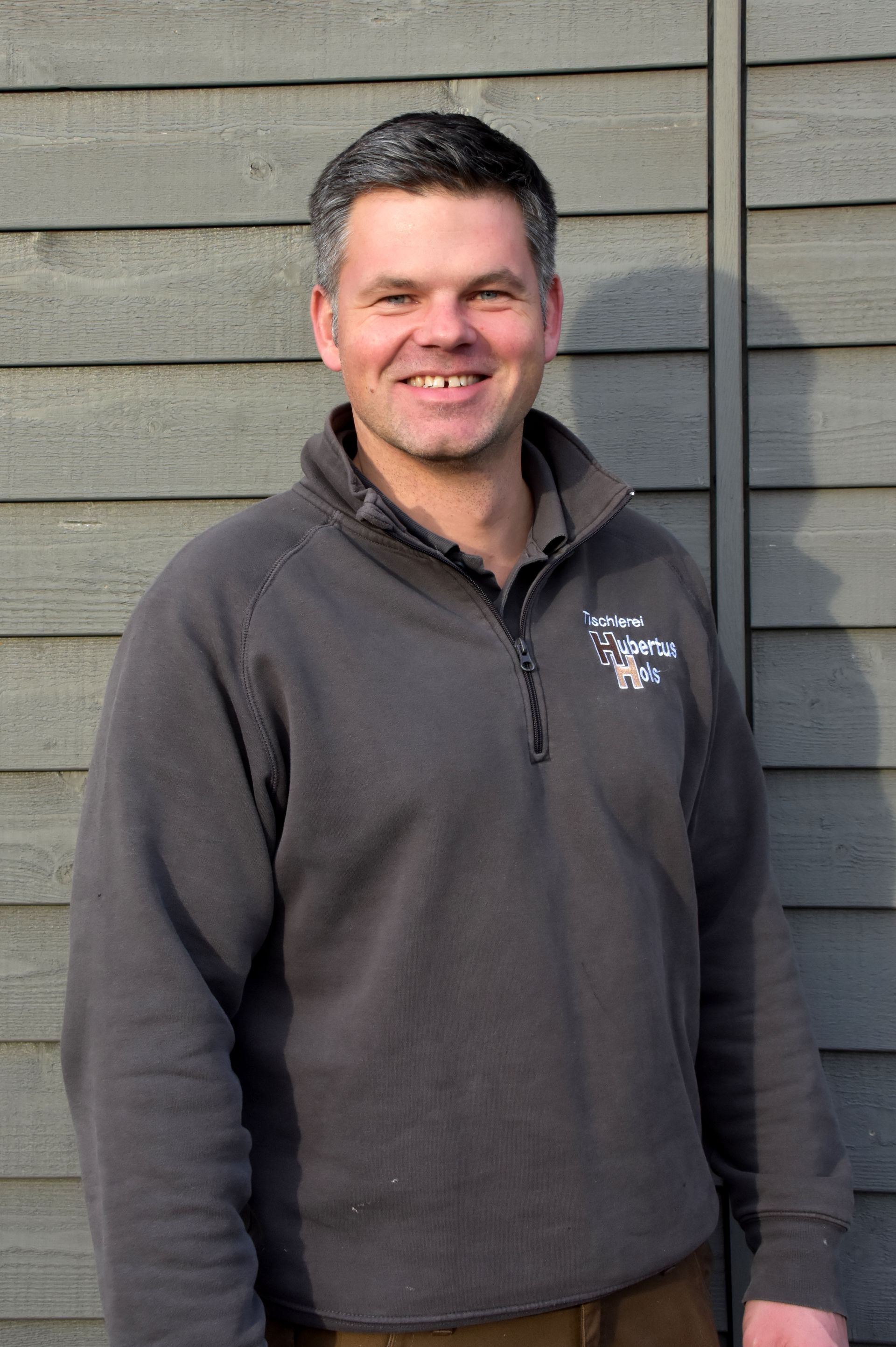 Ein Mann in einem grauen Pullover lächelt vor einer Holzwand.