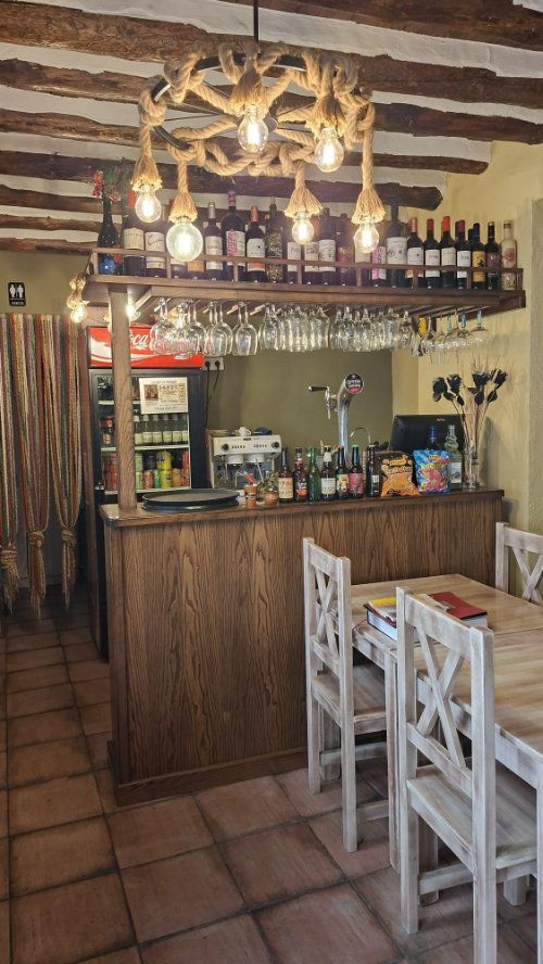 Interior de bar rústico y acogedor con barra de madera, lámparas colgantes y sillas de comedor blancas.