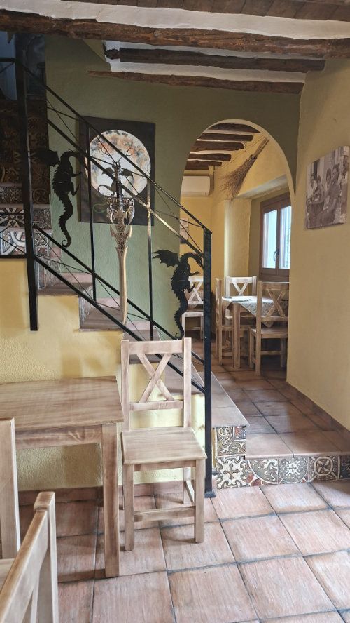Comedor rústico con suelos de baldosas, vigas de madera, puerta arqueada y escalera con barandilla negra.