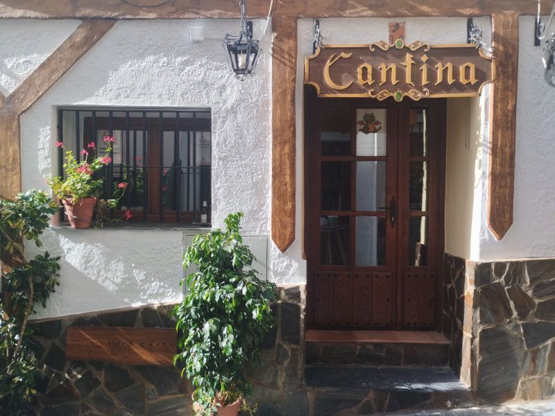 Entrada principal a una cantina con puerta de madera, paredes de estuco blanco y plantas en macetas.