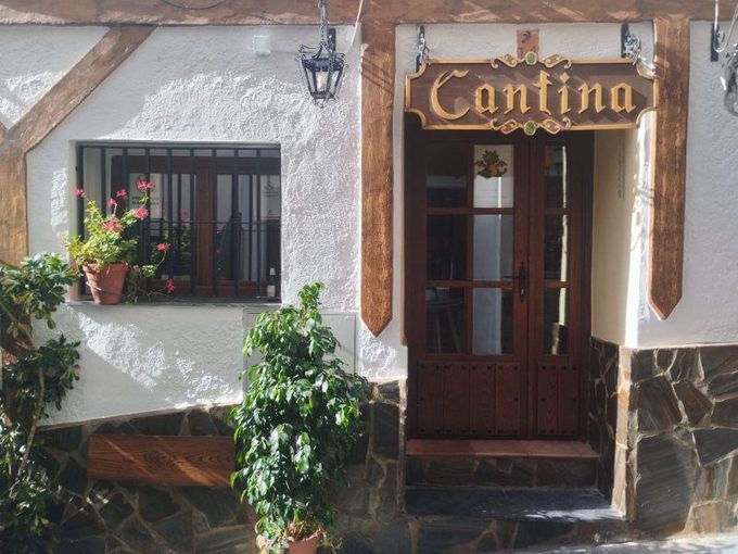 Entrada principal a una cantina con puerta de madera, paredes de estuco blanco y plantas en macetas.