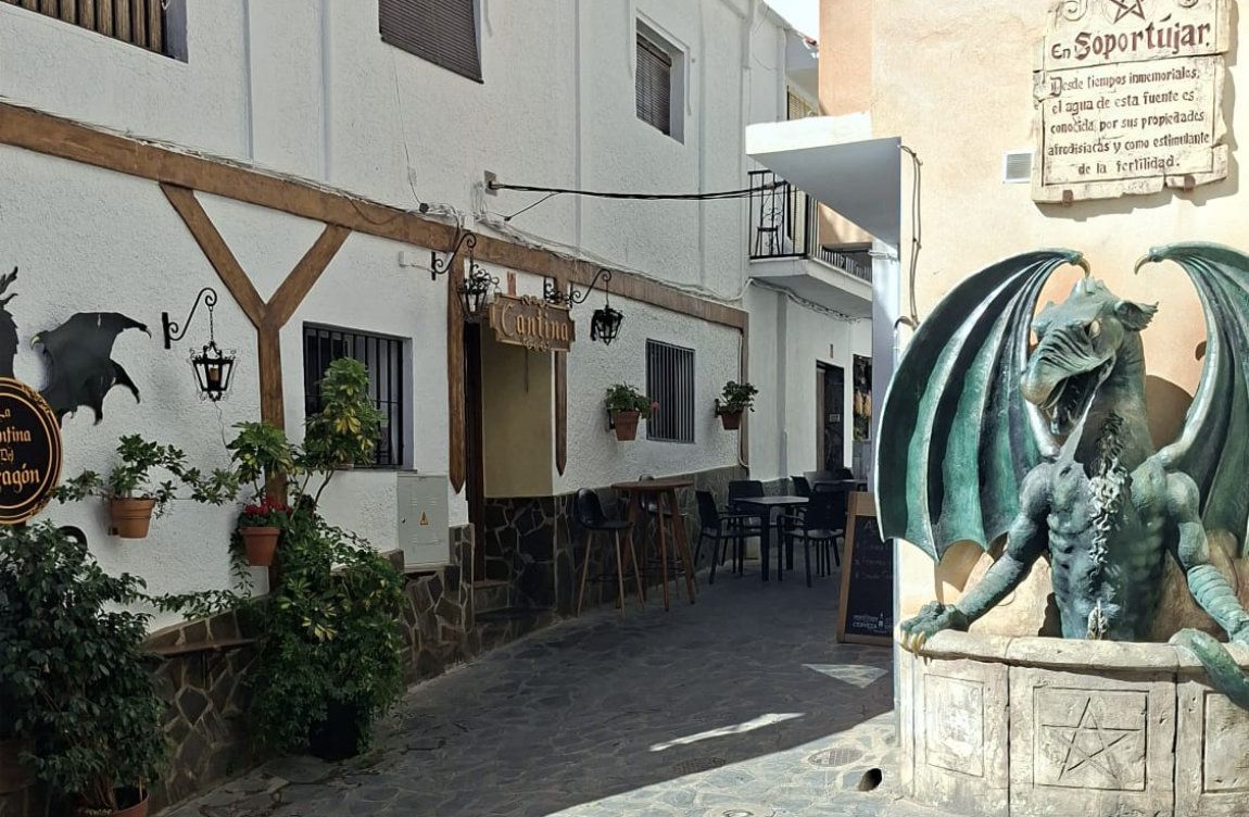 Calle estrecha y blanca con plantas en macetas y una gran estatua de un dragón alado en primer plano.