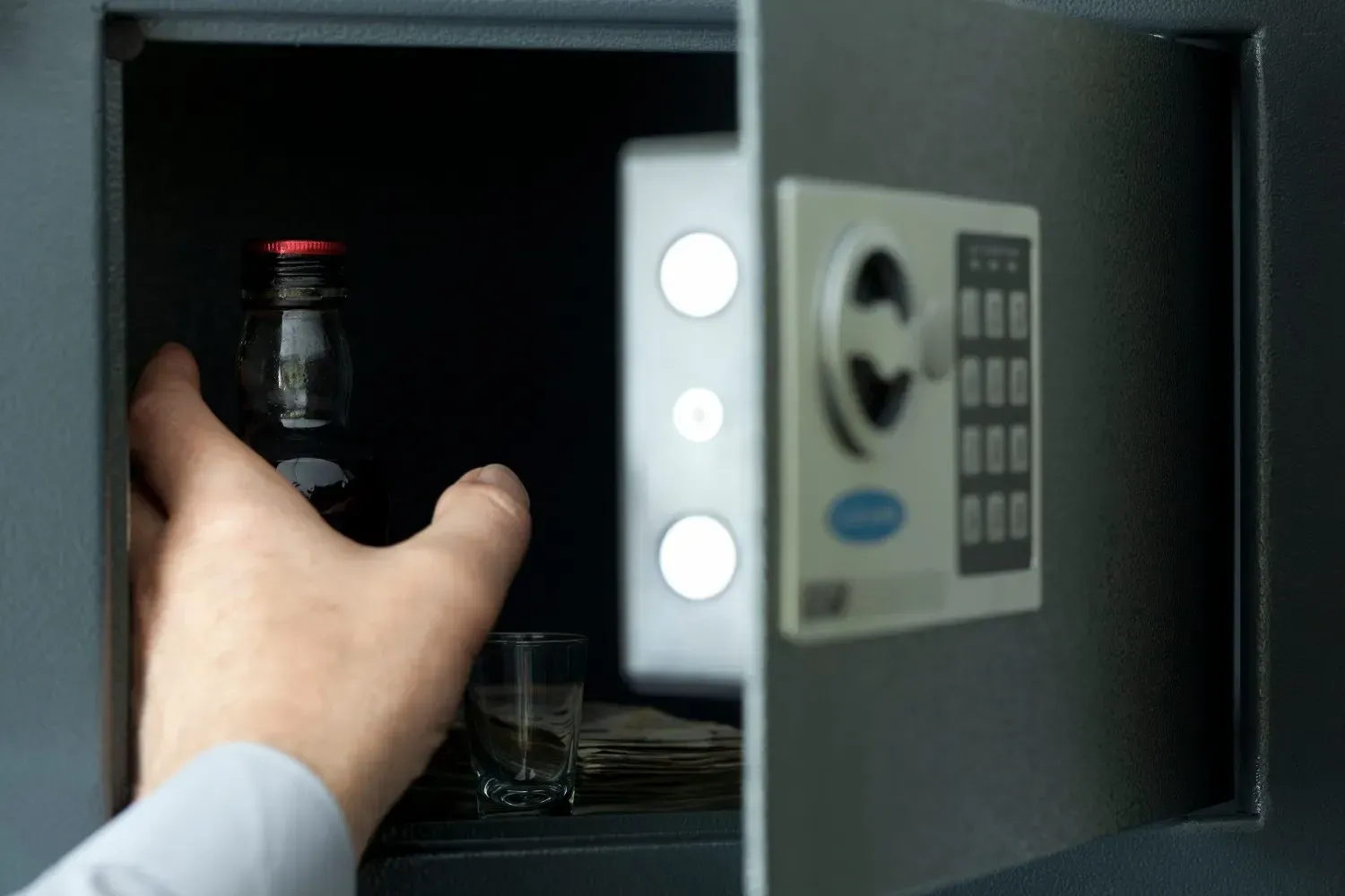 Una persona mete la mano en una caja fuerte oscura dentro de una pared para sacar una botella de vidrio.