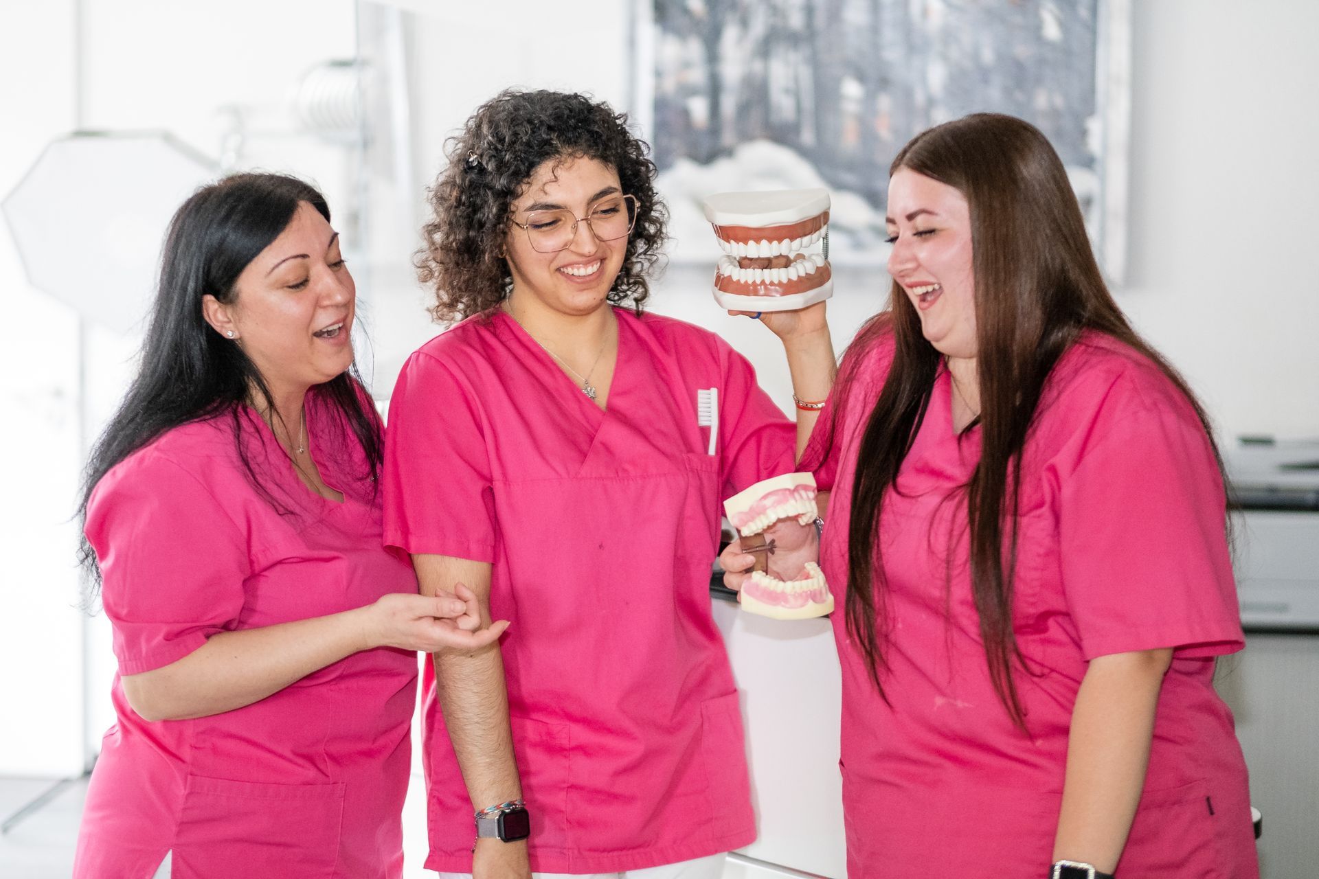 Drei Zahnarzthelferinnen in rosa Kitteln untersuchen lächelnd in einer Praxis ein Zahnmodell.