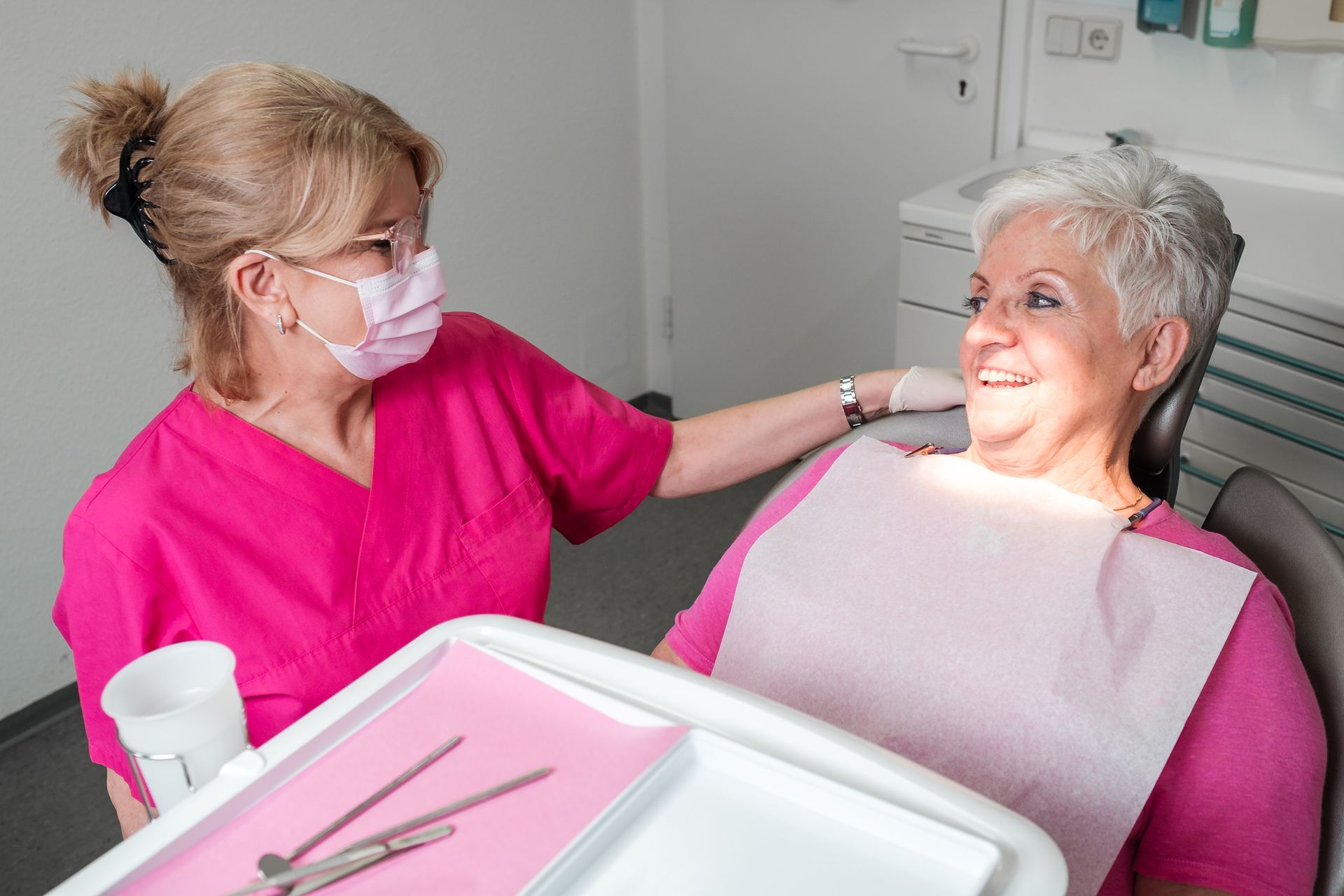 Ein Zahnarzt in rosa OP-Kleidung beruhigt eine lächelnde ältere Frau im Zahnarztstuhl; daneben steht ein Tablett mit Instrumenten.