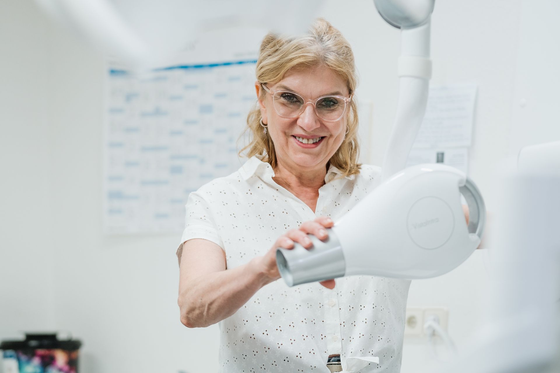 Ein lächelnder Zahnarzt mit Brille hält in einer hell erleuchteten Praxis ein zahnärztliches Röntgengerät.