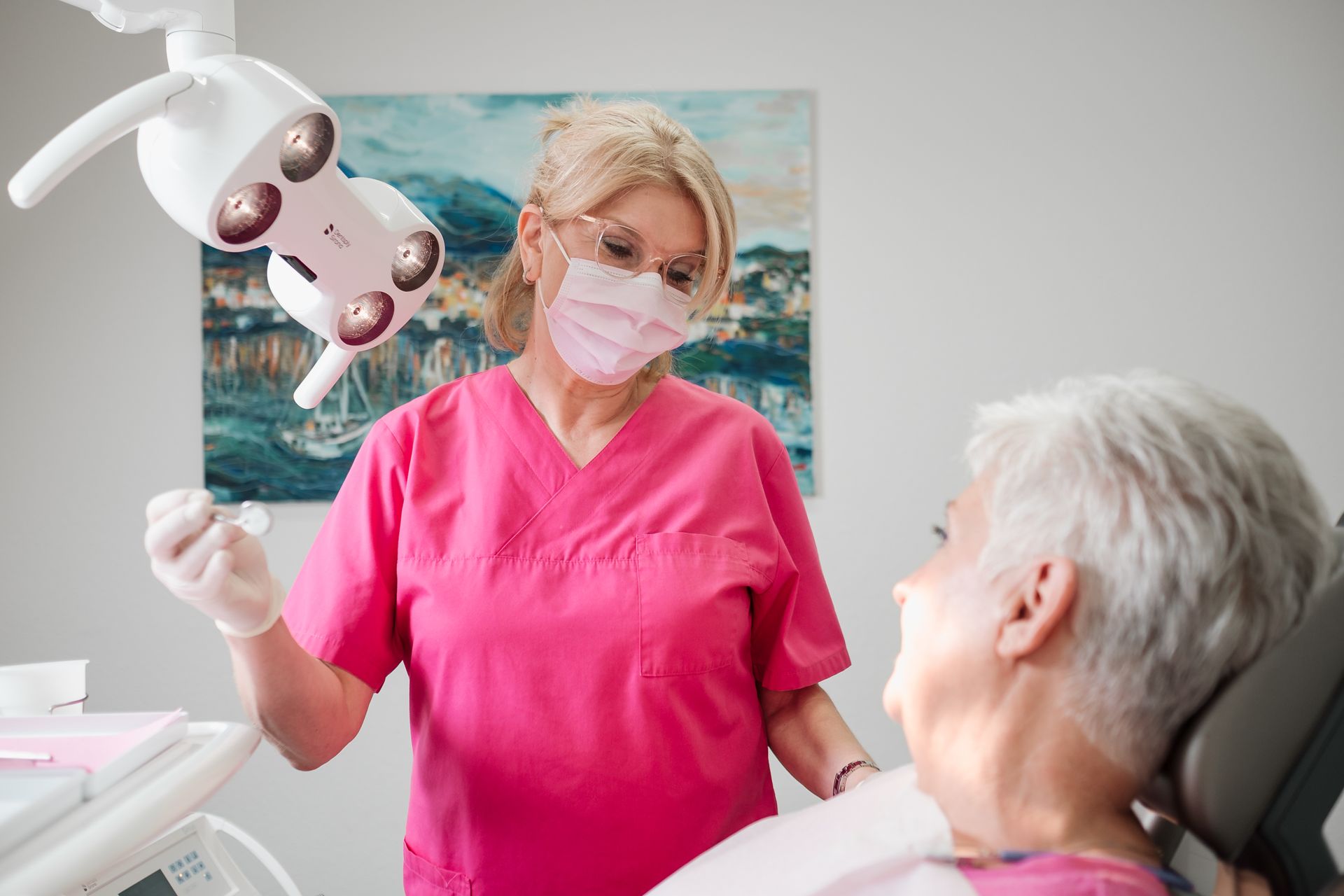 Ein Zahnarzt in rosa OP-Kittel und Maske untersucht den Mund eines Patienten in einem Zahnarztstuhl unter einer hellen Deckenlampe.