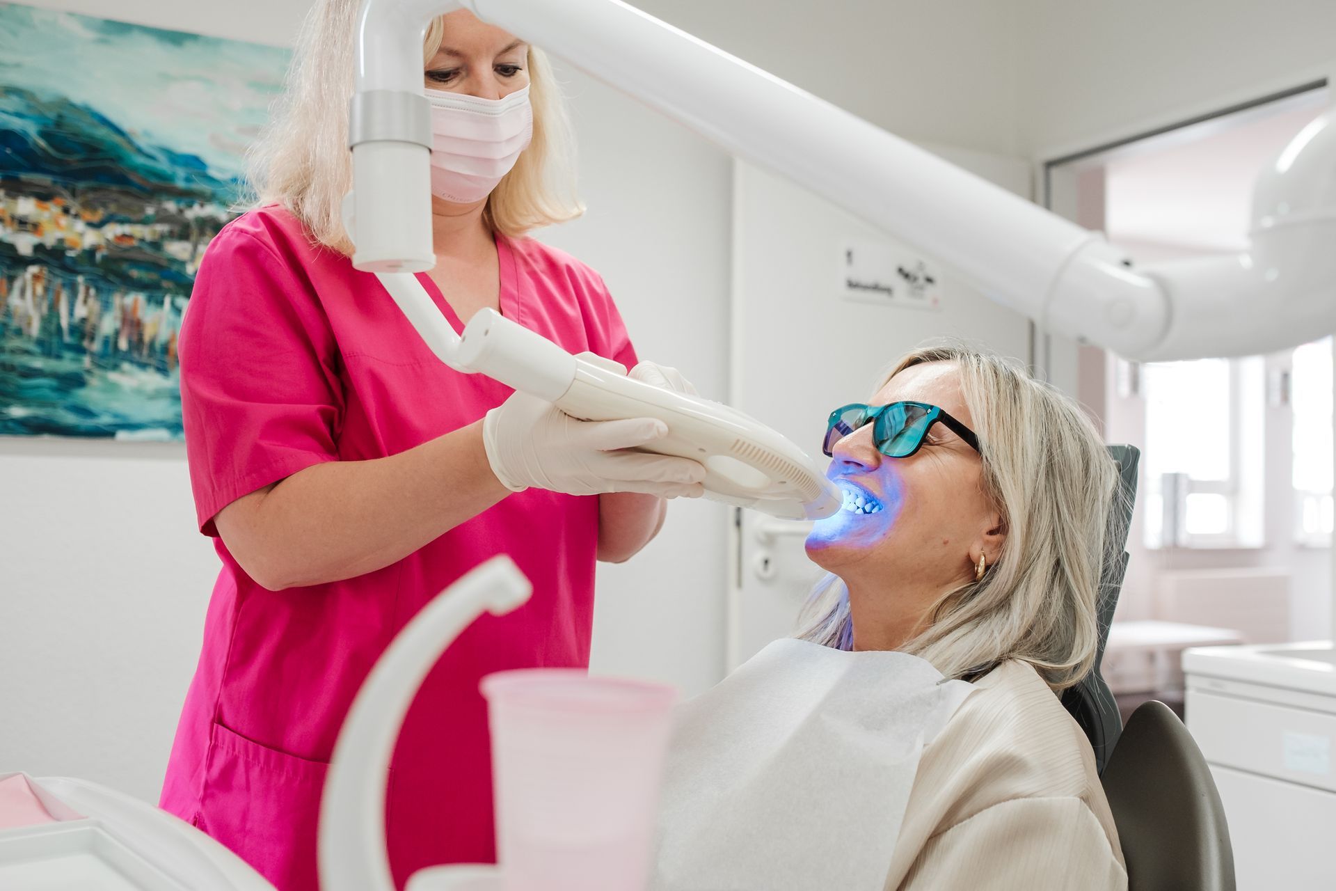 Zahnarzt in rosa OP-Kittel und Maske verwendet ein Blaulichtgerät an den Zähnen eines Patienten. Der Patient trägt eine Brille und ein Lätzchen.
