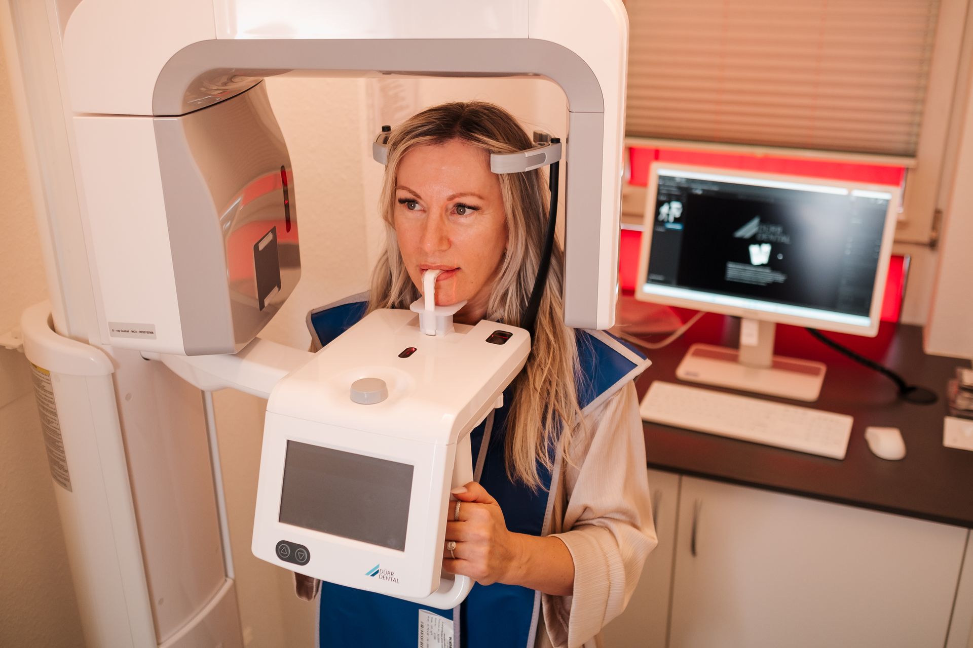 Frau unterzieht sich einer zahnärztlichen Röntgenuntersuchung. Sie hält das Gerät fest und blickt nach vorne. Die Untersuchung findet in einer Zahnarztpraxis statt.