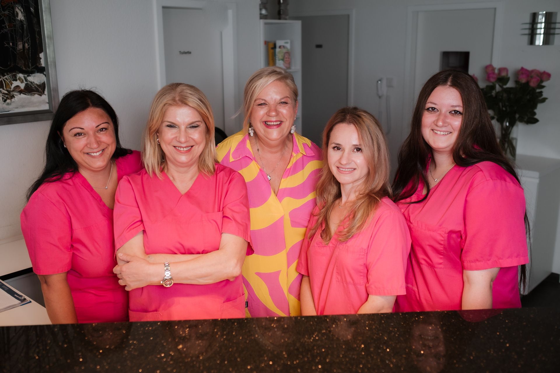 Fünf Frauen, meist in rosa OP-Kleidung, lächeln hinter einem Empfangstresen. Eine Frau trägt ein gelb-rosa gemustertes Hemd.