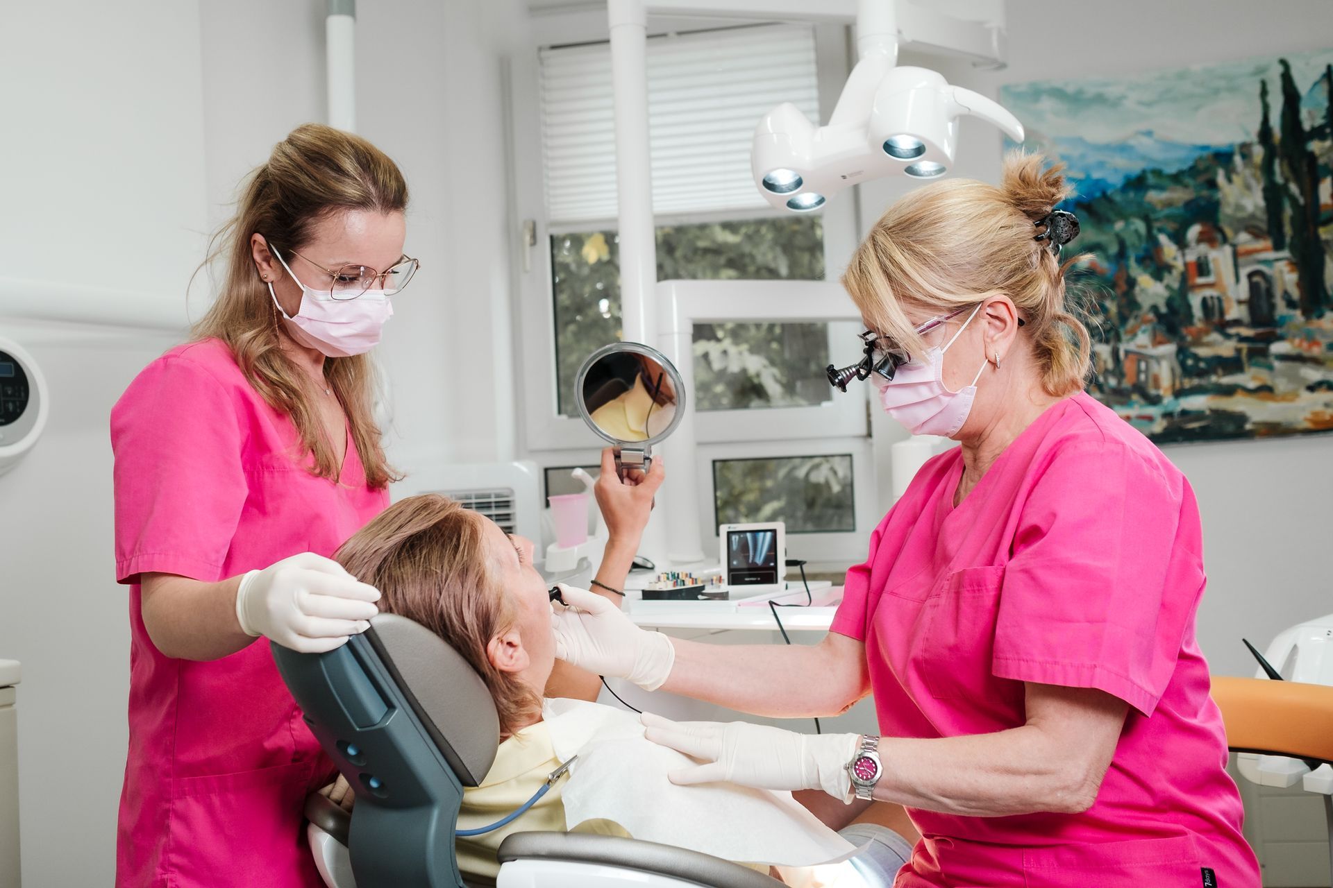 Zwei Zahnärzte untersuchen in einer hellen Praxis den Mund eines Patienten. Beide tragen rosa Kittel, Mundschutz und Handschuhe. Einer hält einen Spiegel.