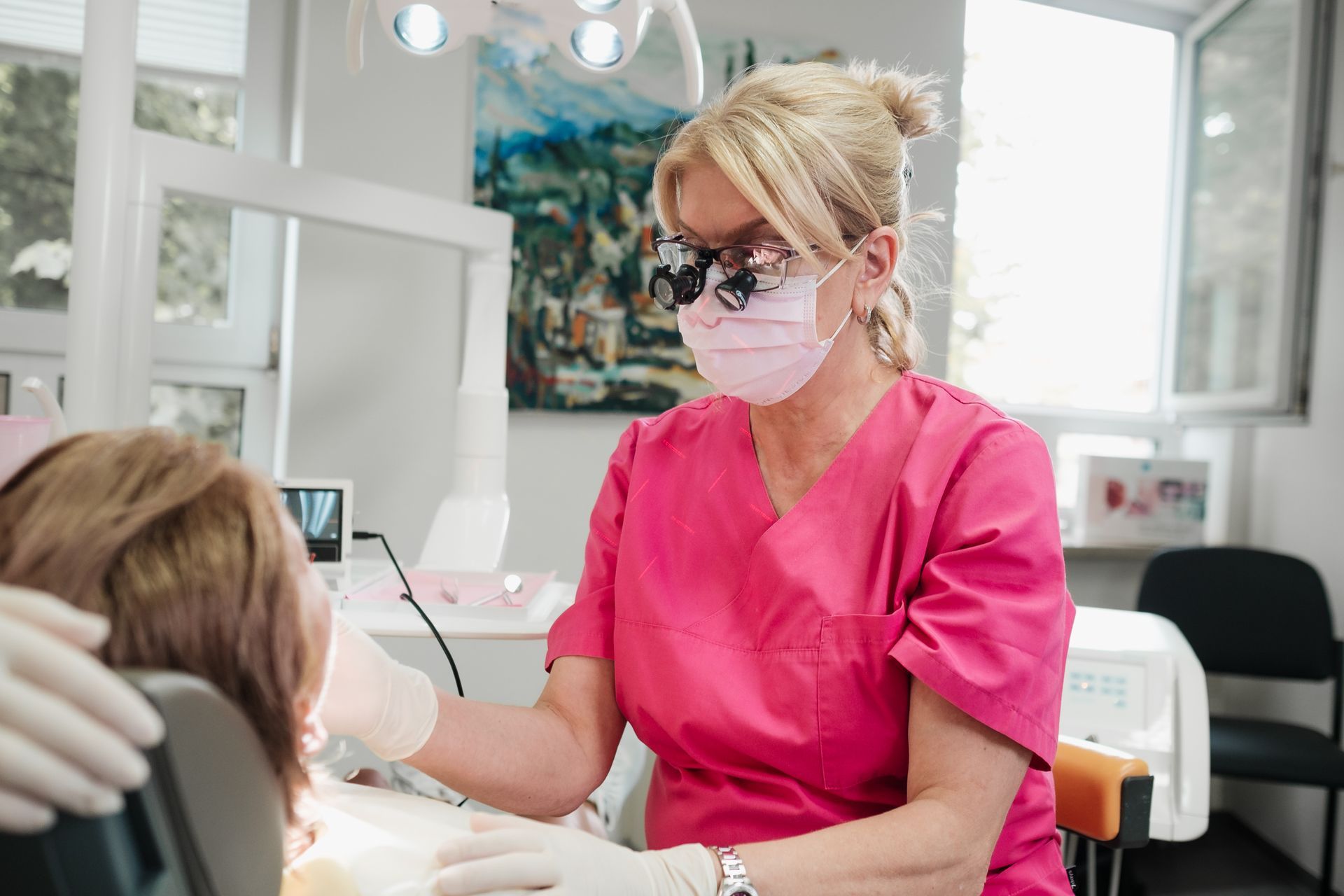 Ein Zahnarzt in rosa OP-Kleidung und mit Lupen untersucht in einer Zahnarztpraxis den Mund eines Patienten.