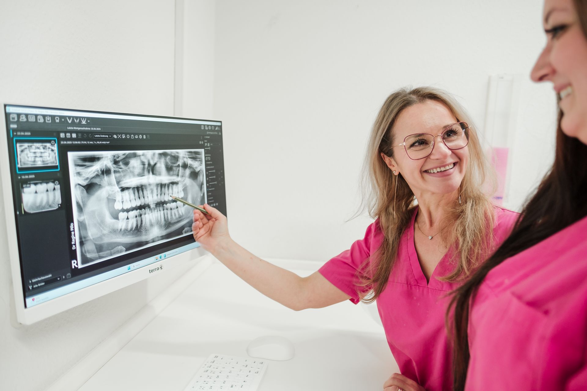 Zwei Zahnärzte in rosa Kitteln betrachten ein Zahnröntgenbild auf einem Computerbildschirm; einer zeigt lächelnd auf das Röntgenbild.