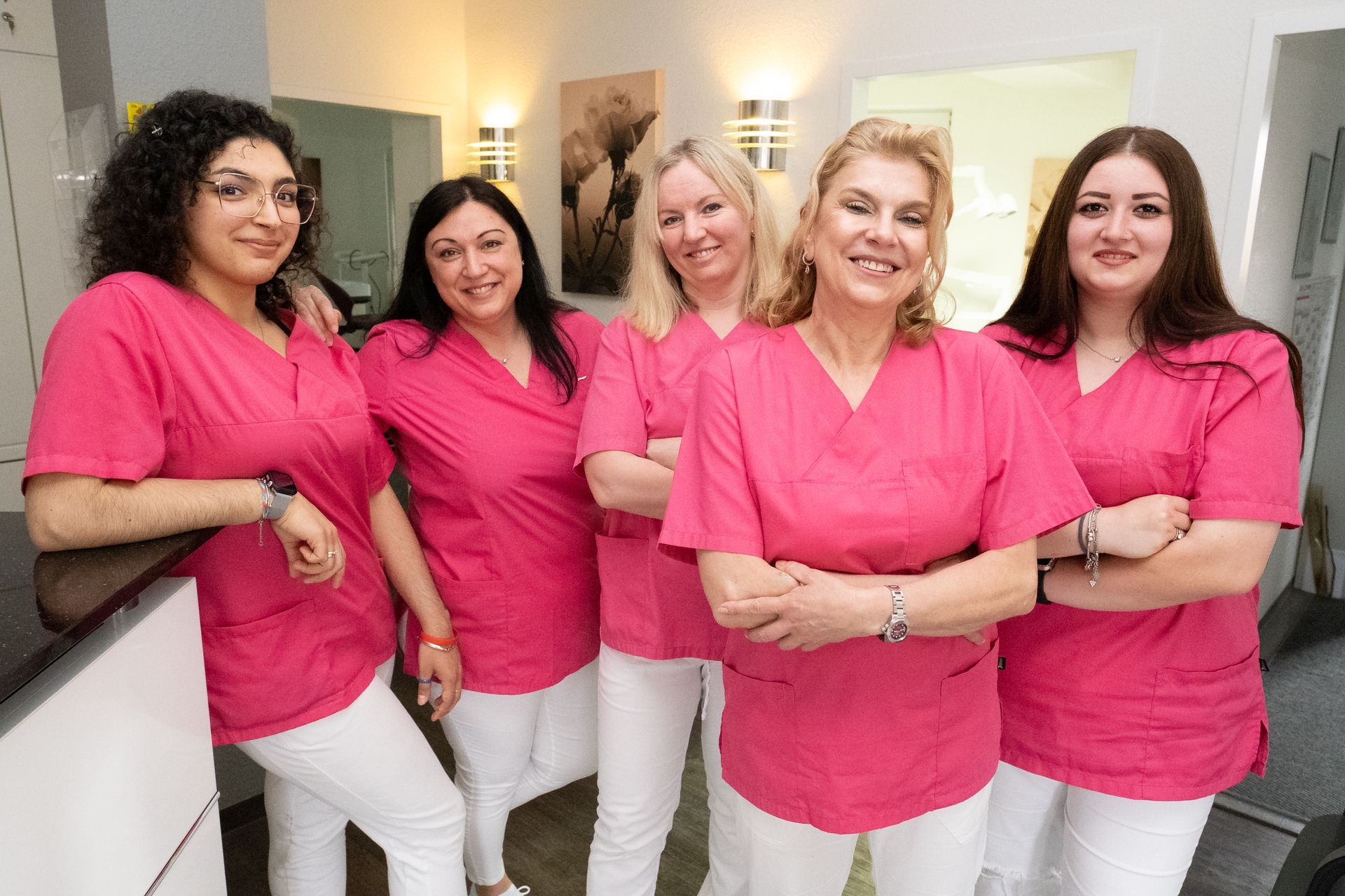 Fünf Frauen in rosa OP-Kleidung und weißen Hosen, wahrscheinlich medizinisches Fachpersonal, lächeln in einem Büro.