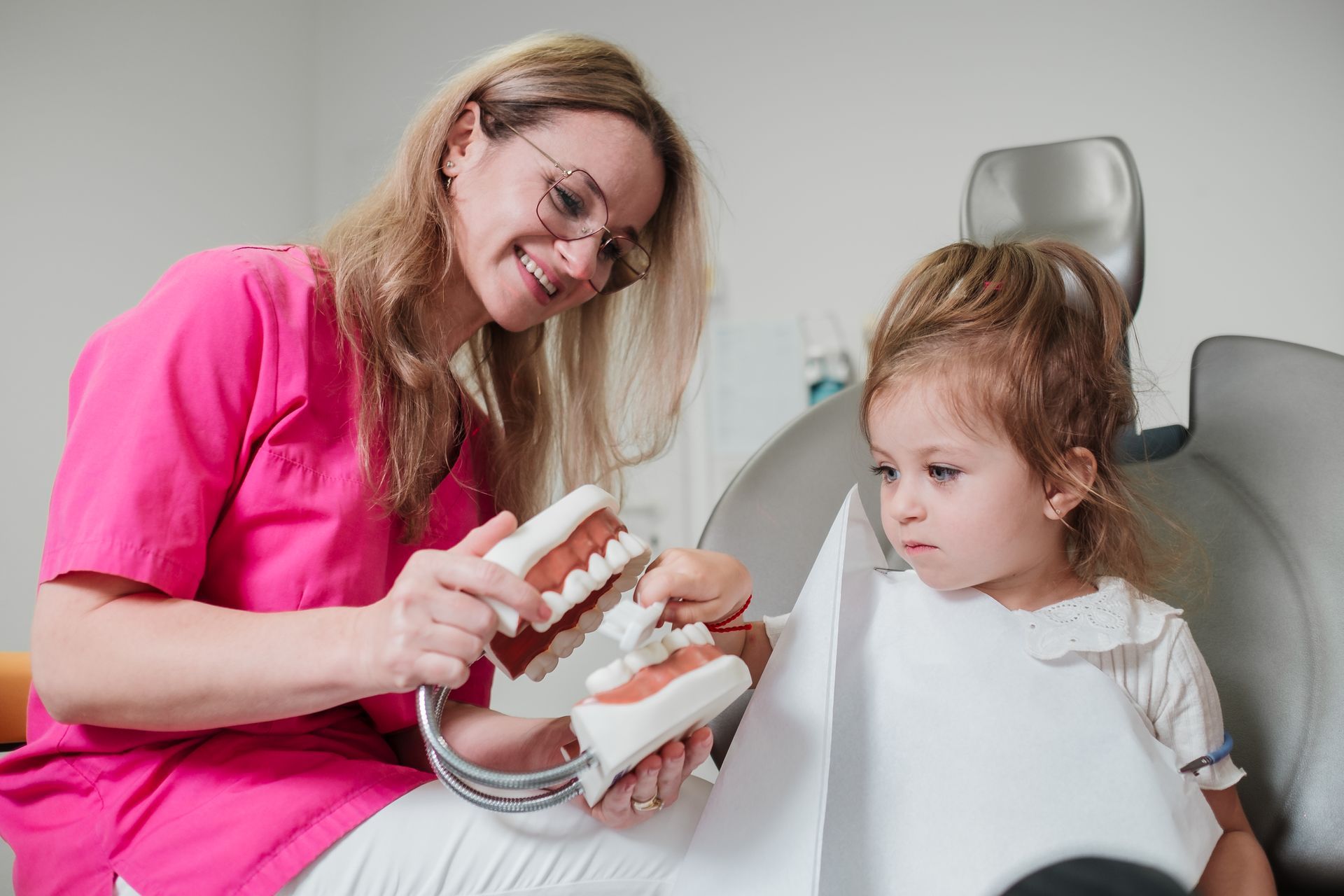 Ein Zahnarzt in einer rosa Uniform erklärt einem jungen Mädchen im Zahnarztstuhl anhand eines Modells die Zähne.