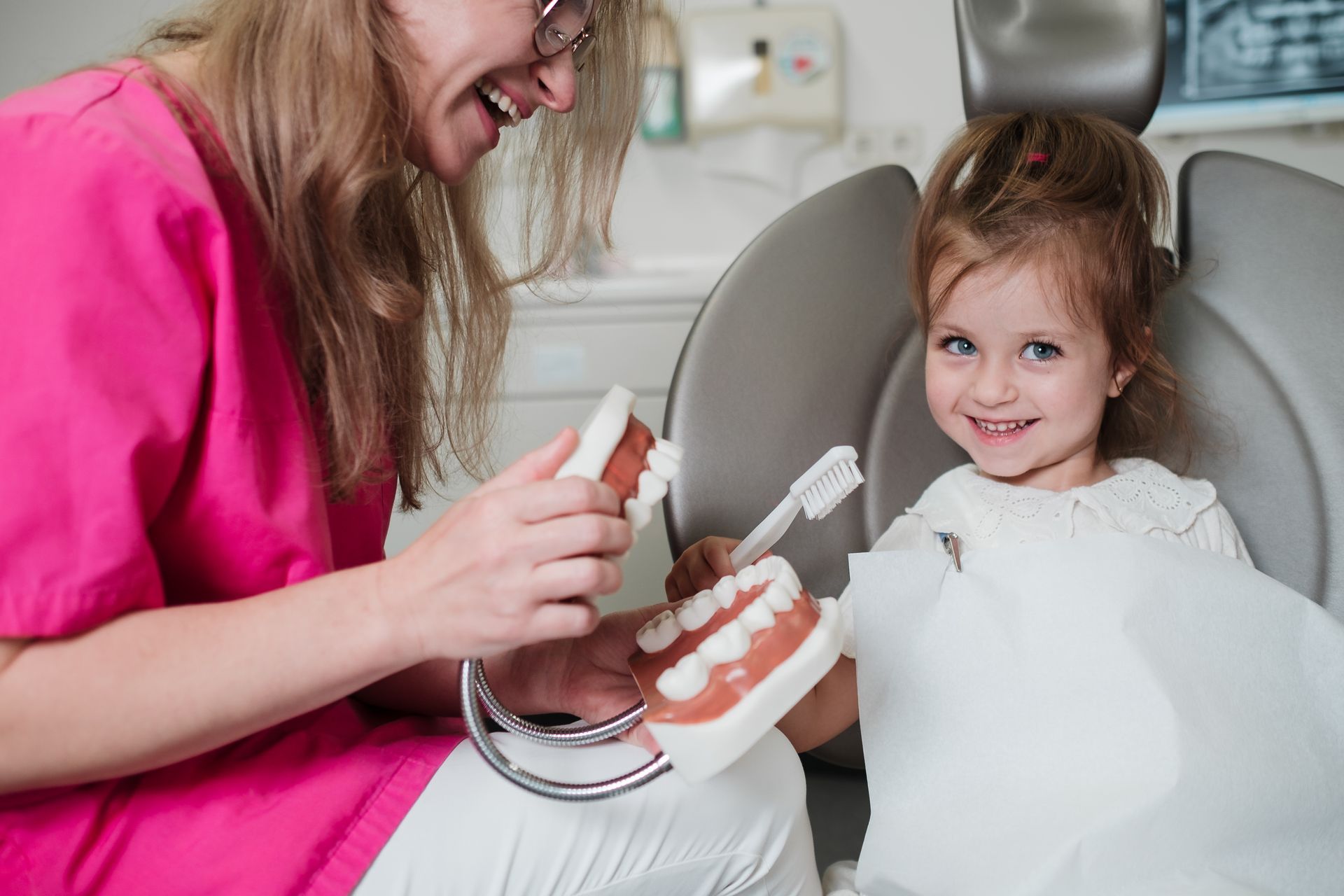Zahnarzt zeigt einem jungen Mädchen in einem Zahnarztstuhl ein Zahnmodell und eine Zahnbürste. Beide lächeln.