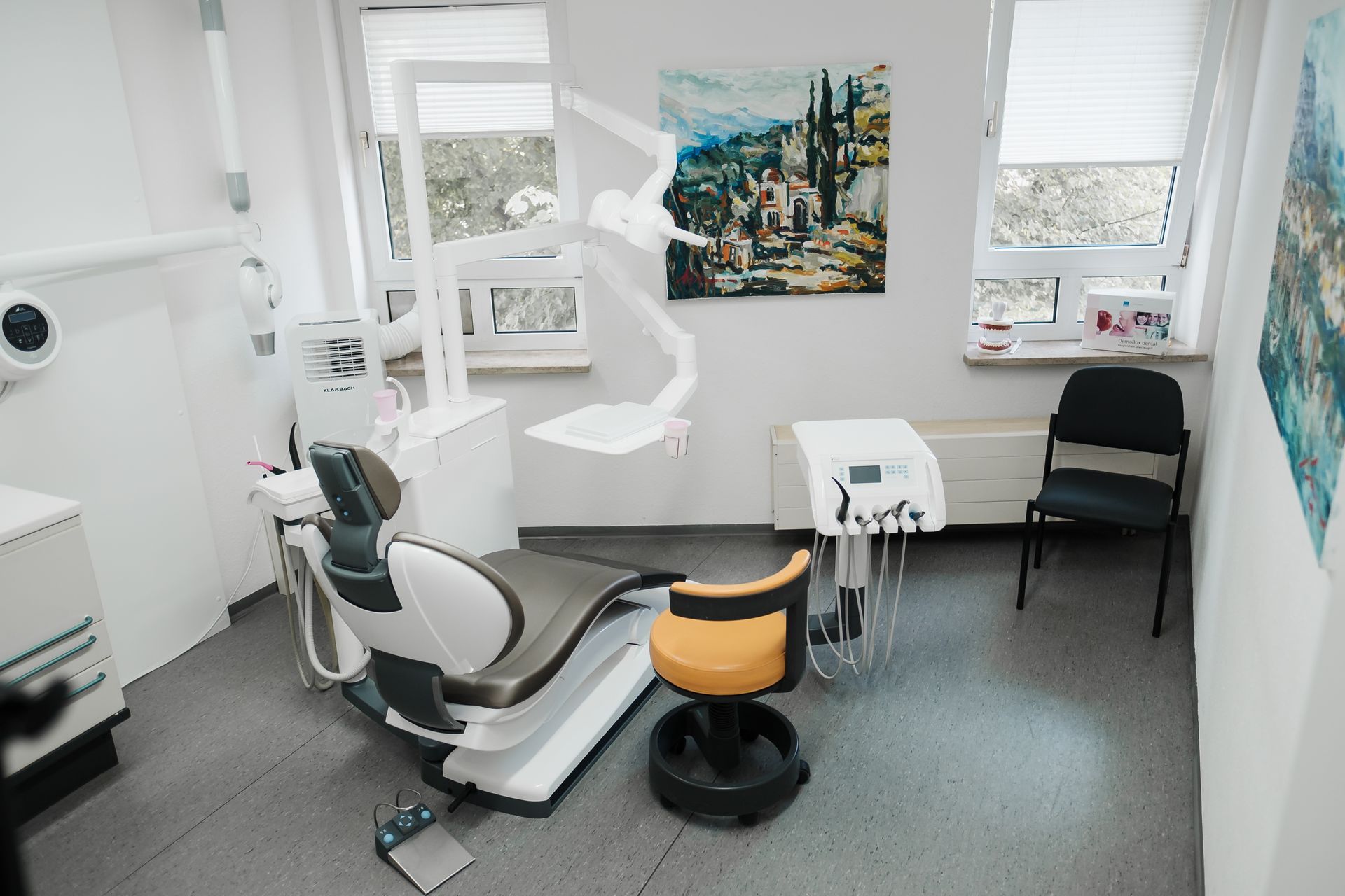 Eine Zahnarztpraxis mit Untersuchungsstuhl, Geräten, Kunstwerken und einem schwarzen Stuhl. Der Raum ist hell erleuchtet und wird durch zwei Fenster mit natürlichem Licht beleuchtet.