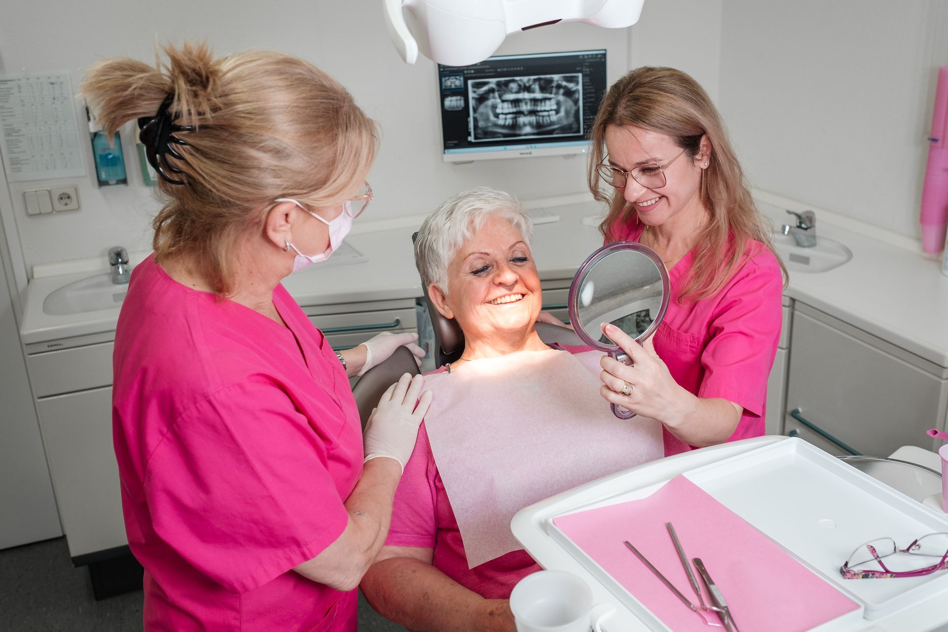 Zahnarztpatientin schaut lächelnd in den Spiegel. Zwei Zahnärztinnen in rosa Kitteln assistieren in einer Zahnarztpraxis.