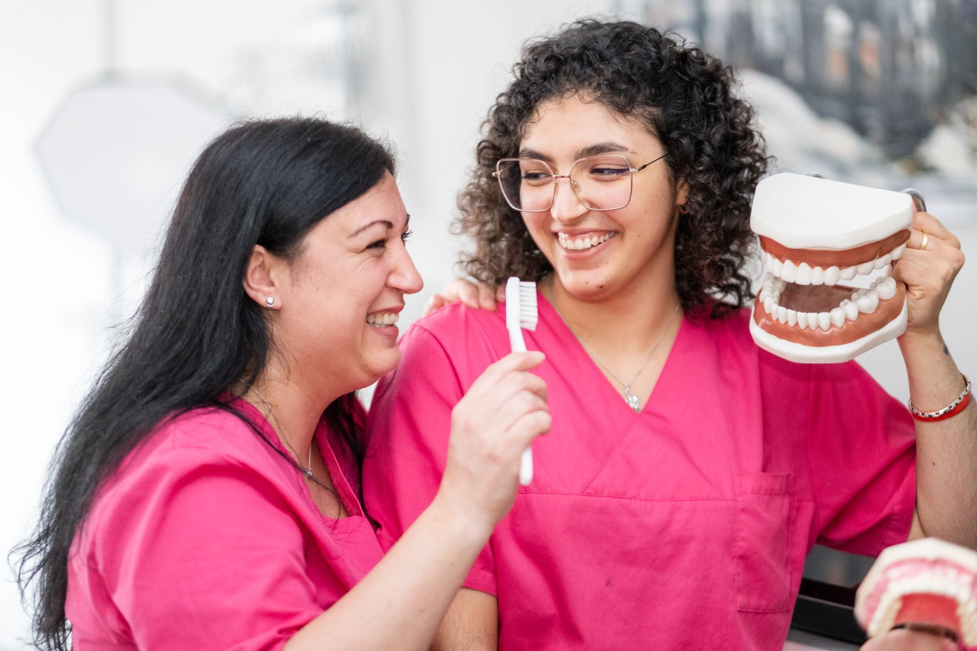 Zwei lächelnde Zahnärzte in rosa Kitteln, einer hält eine Zahnbürste, der andere ein Zahnmodell.