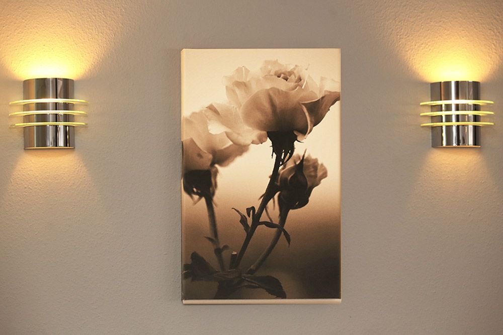 Zwei Wandlampen beleuchten ein sepiafarbenes Rosengemälde an einer grauen Wand.
