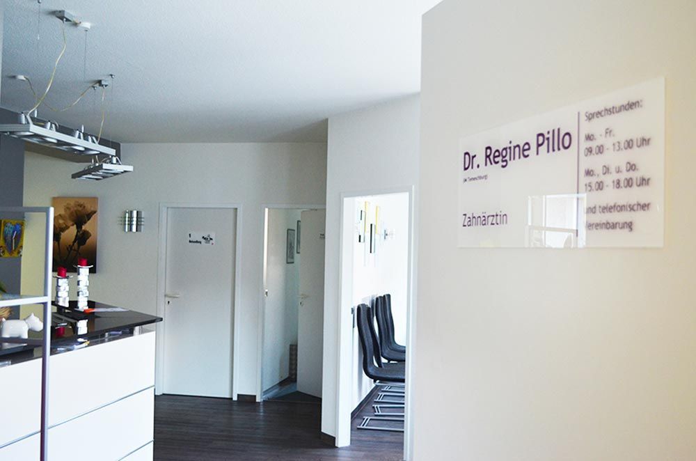 Der Empfangsbereich einer Zahnarztpraxis mit einem Schild mit den Sprechzeiten. Weiße Wände, Türen und Möbel.