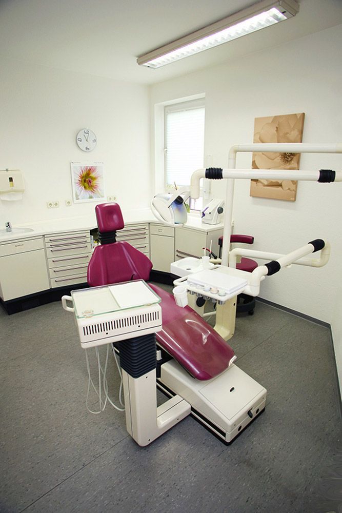 Ein Zahnarztzimmer mit einem rosa-weißen Stuhl, Instrumenten und Schränken. Von der Decke strahlt helles Licht.