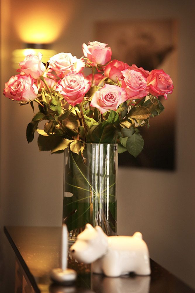 Rosa Rosen in einer durchsichtigen Vase stehen auf einer dunklen Fläche mit einer kleinen weißen Hundefigur. Sanftes Licht schafft eine warme Atmosphäre.