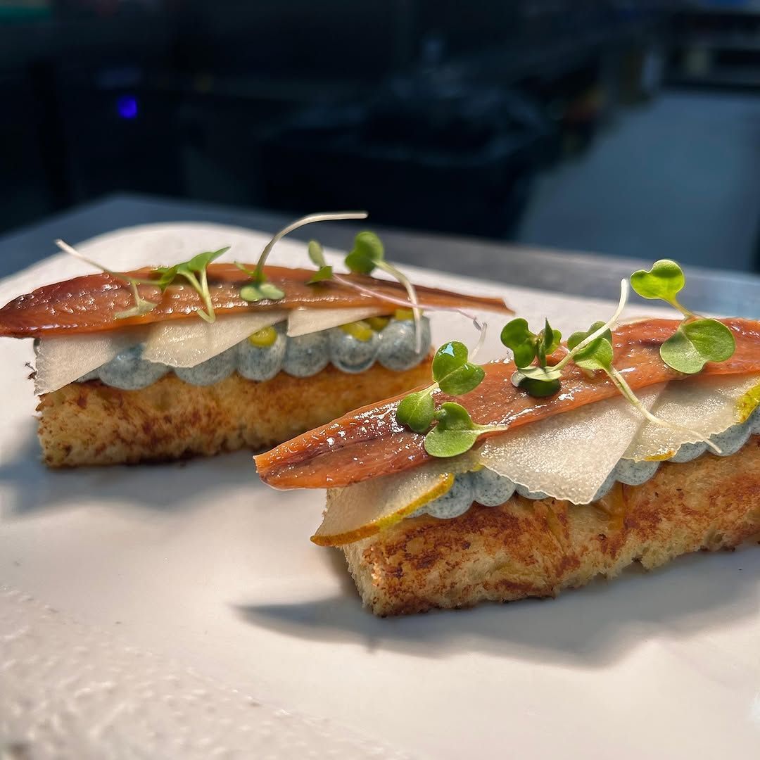 Dos elegantes canapés estilo tartine con pescado, crema y microvegetales sobre un plato blanco.