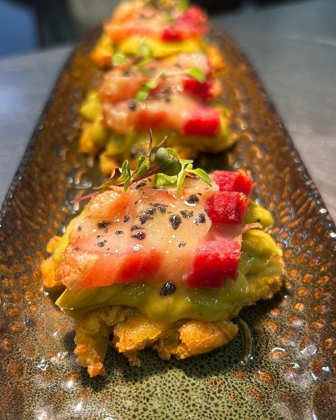 Bocaditos de tartar de aguacate y salmón en un plato negro texturizado, adornados con semillas de sésamo y microvegetales.