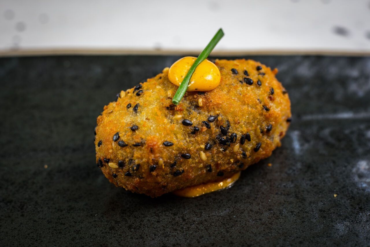 Croqueta frita de color marrón dorado, cubierta con una guarnición verde sobre un plato oscuro.