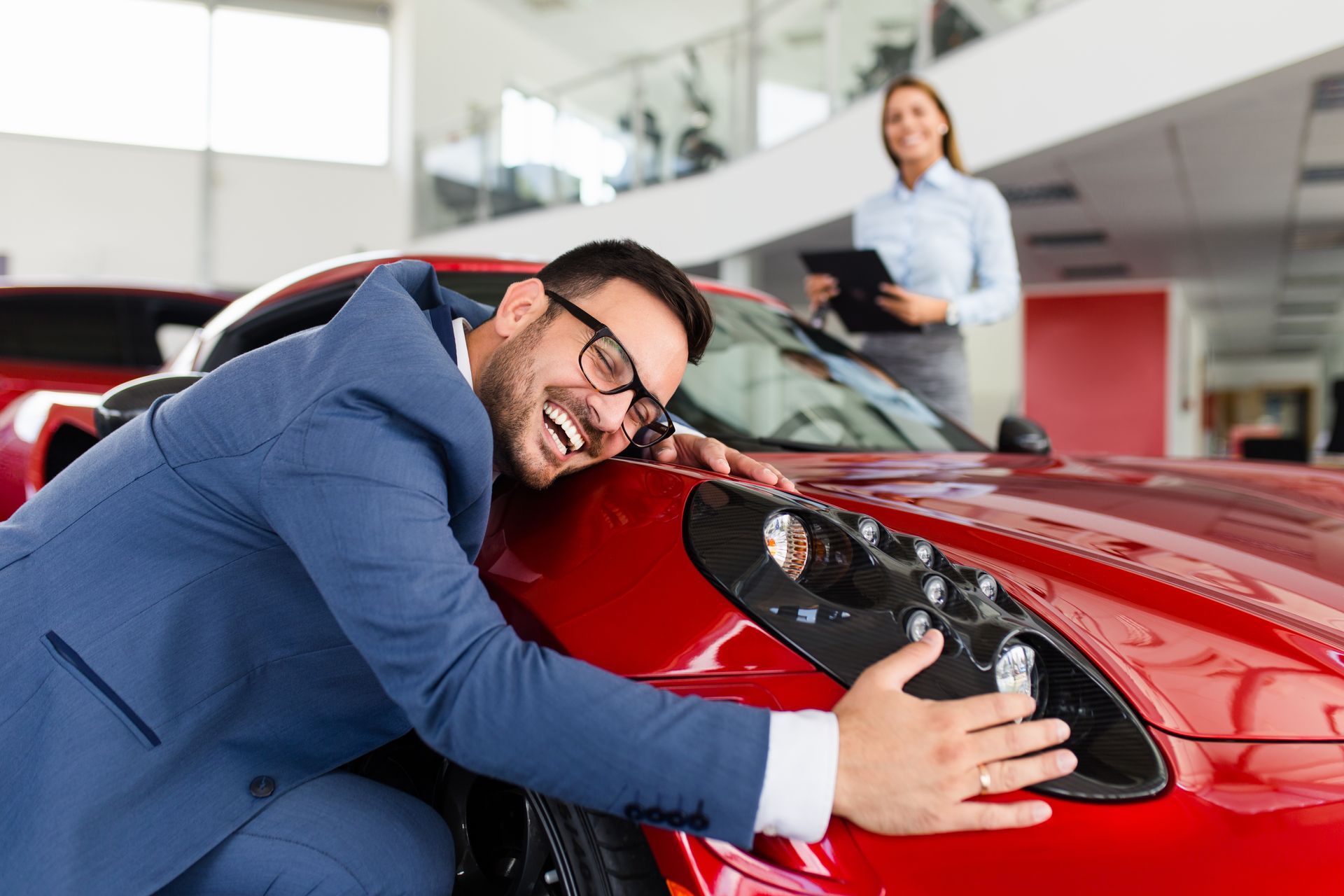 Ein Mann umarmt lächelnd einen roten Sportwagen in einem Ausstellungsraum. Im Hintergrund schaut eine Frau zu.