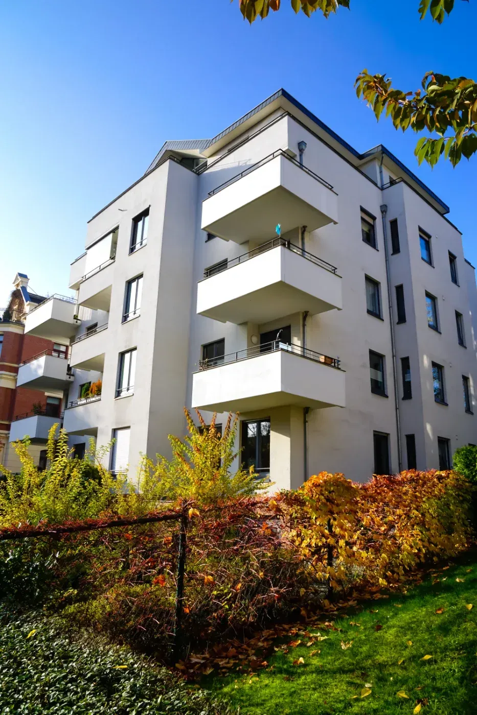 Mehrstöckiges Wohnhaus mit weißen Balkonen, umgeben von Herbstlaub.
