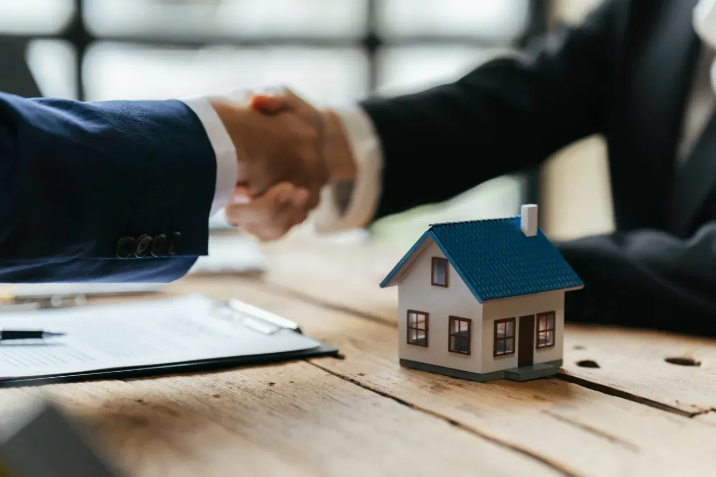 Zwei Personen schütteln sich über einem Miniaturhaus, Dokumenten und einem Stift auf einem Holztisch die Hände.